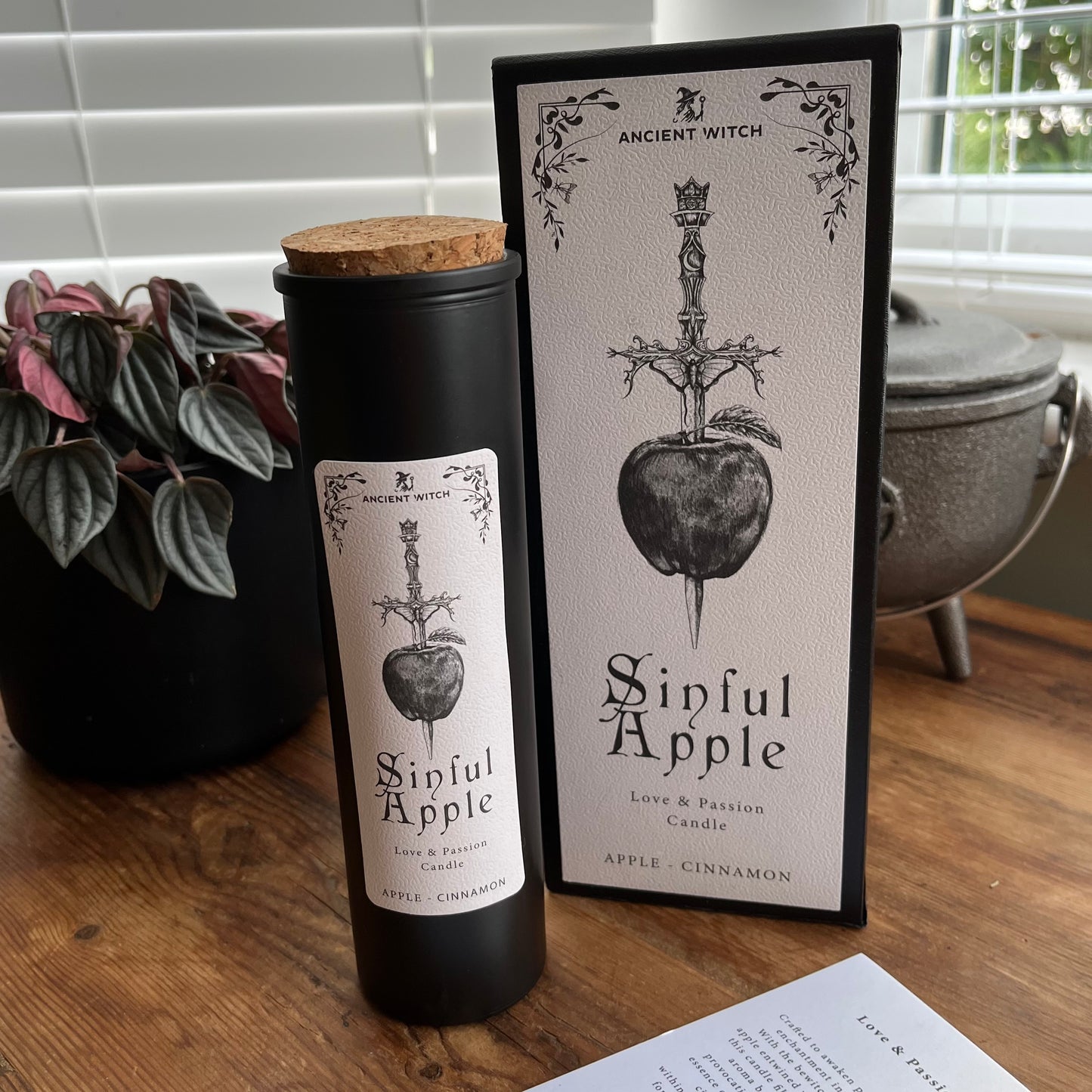 A black cylindrical candle jar with a cork lid stands beside its matching rectangular box on a wooden table. The label and box both show a black-and-white illustration of a sword piercing an apple, with the text 'Ancient Witch' and 'Sinful Apple.