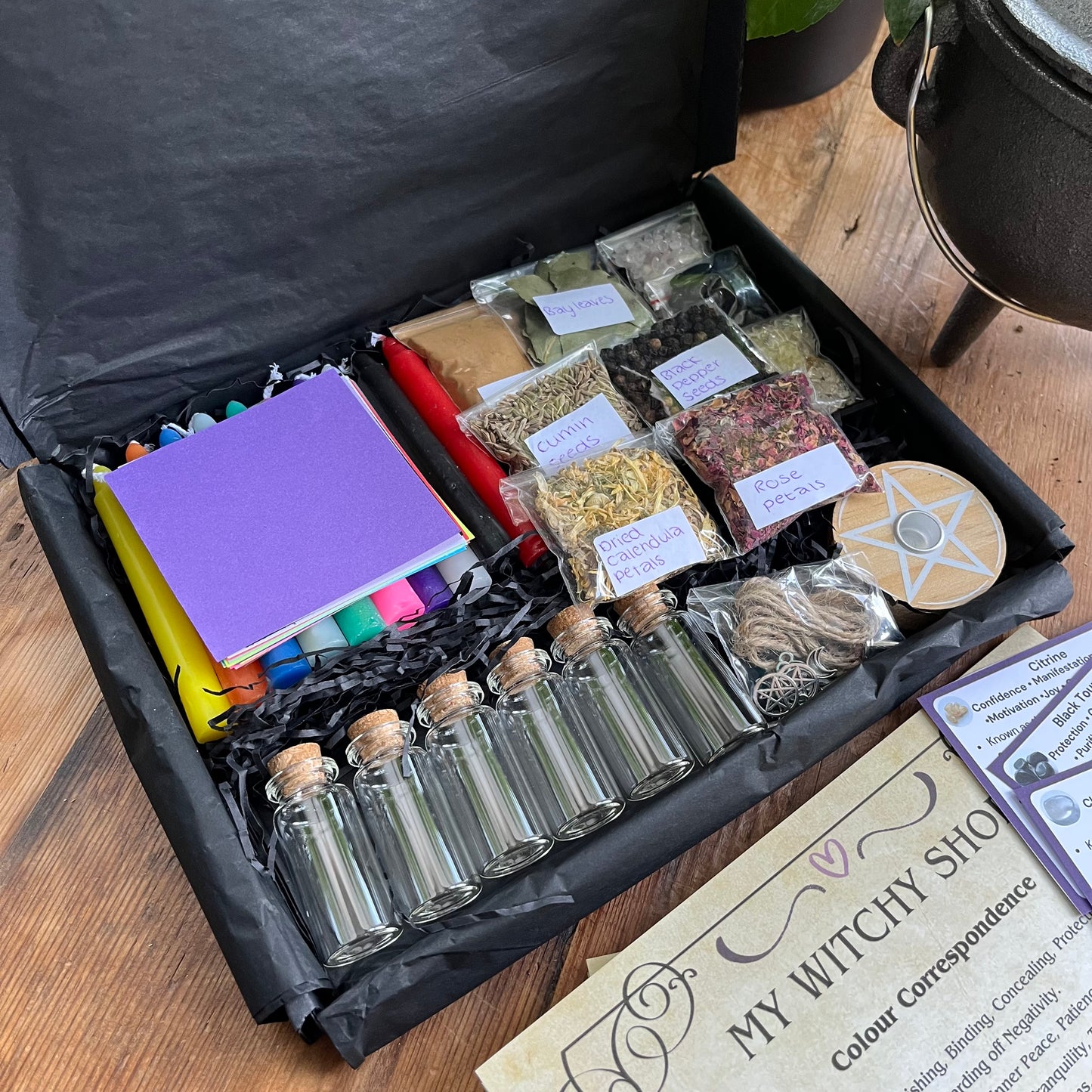 Witchcraft supplies including bottles, herbs, crystals and candles in a black box on a wooden surface.