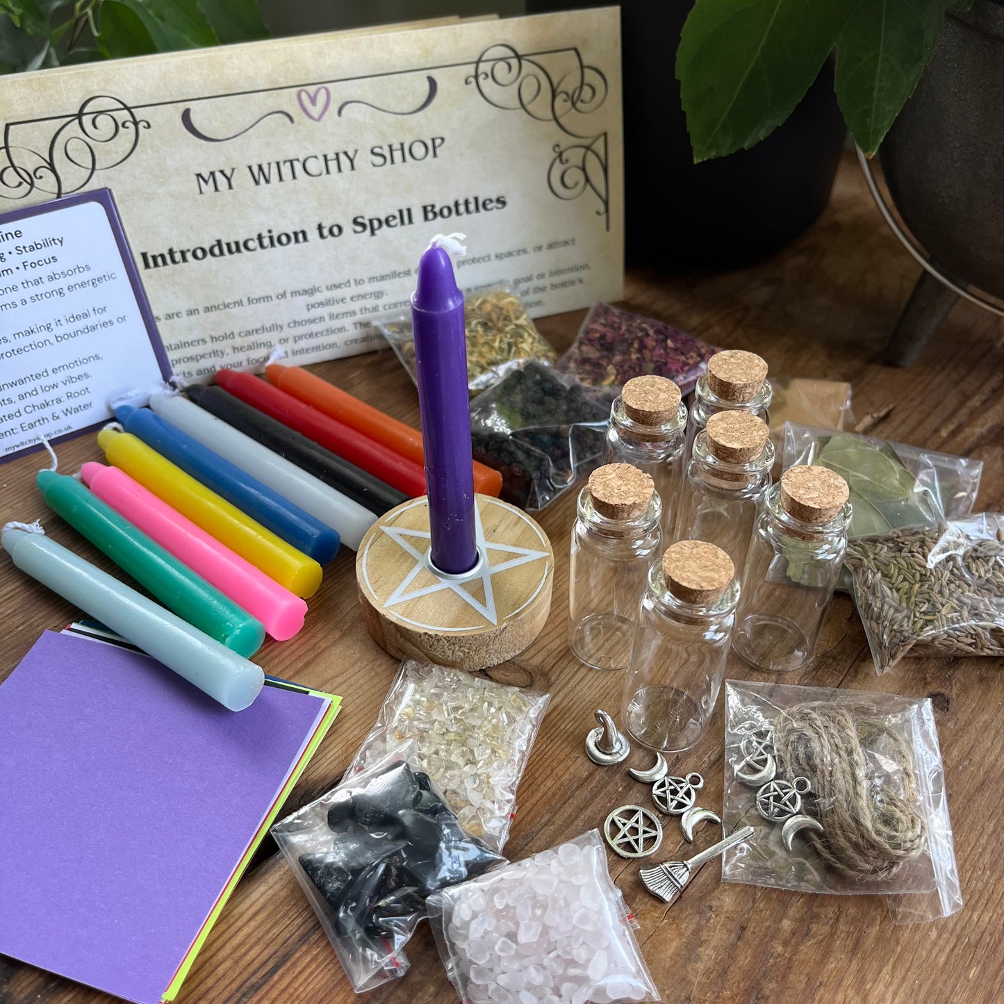 Witchcraft supplies including colored candles, bottles, and a purple candle on a wooden surface with 'My Witchy Shop' introduction book.