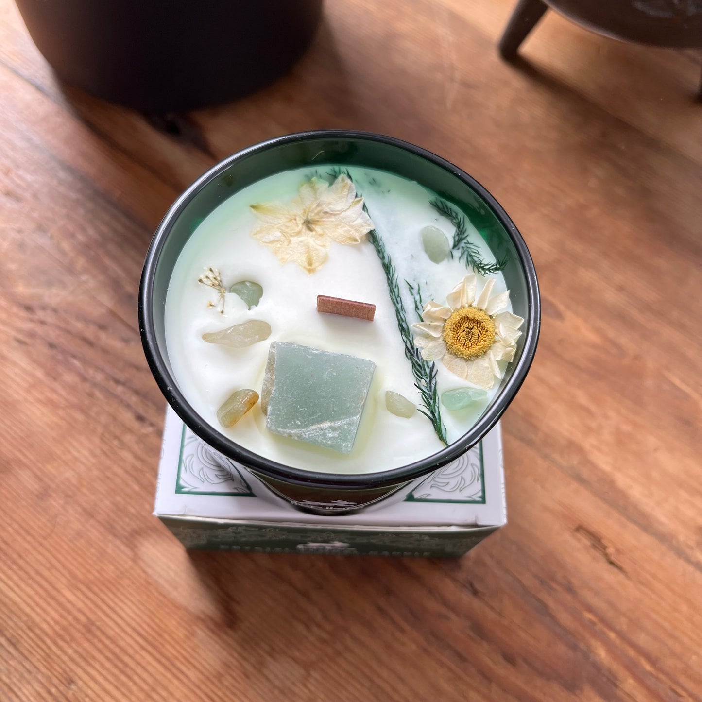 Candle in a decorative container with flowers and herbs on a wooden surface