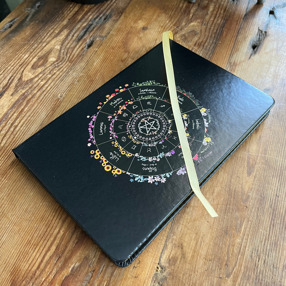 A black hardcover journal with a colorful astrological wheel design on the cover, resting on a rustic wooden table. A light beige ribbon bookmark is tucked inside the cover.