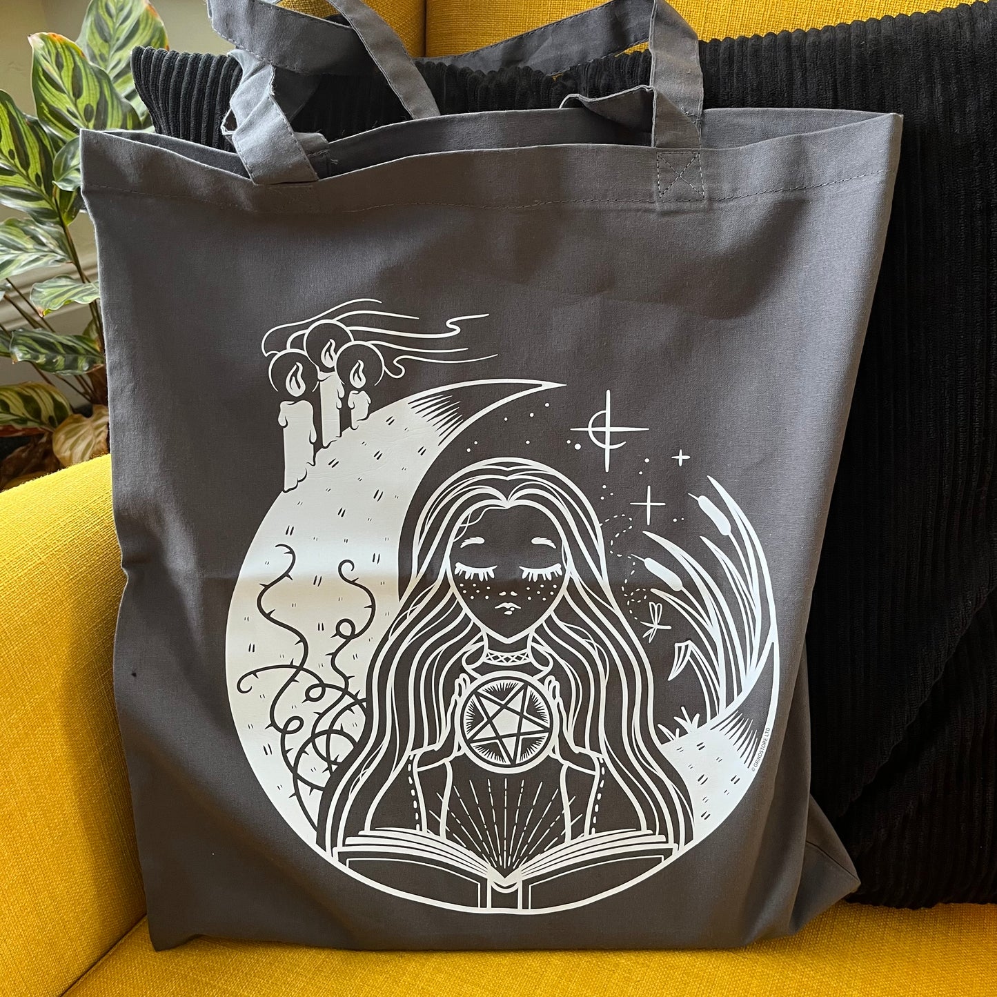 Gray tote bag with white graphic design of a woman with long hair and a pentagram symbol, placed on a yellow surface.