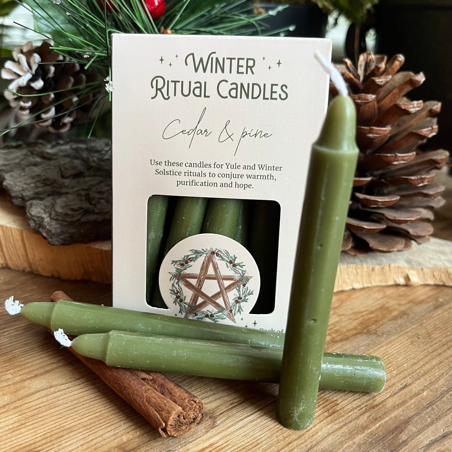 A set of 12 green spell cnadles in a cream coloured box adorned with a pentagram and a holly wreath. Three candles are positioned out of the box and there are yule symbols in the back ground