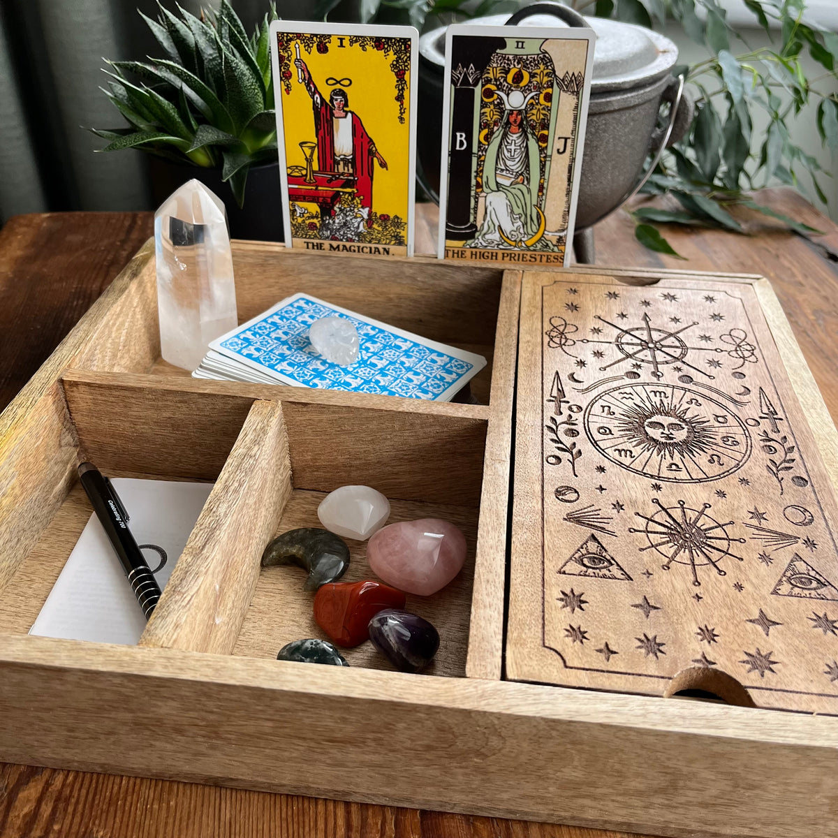 A wooden compartment tray set for tarot card practice, shown open with a crystal point, card deck, tarot cards, polished stones, and a printed wooden box lid on a wooden table with plants in the background.
