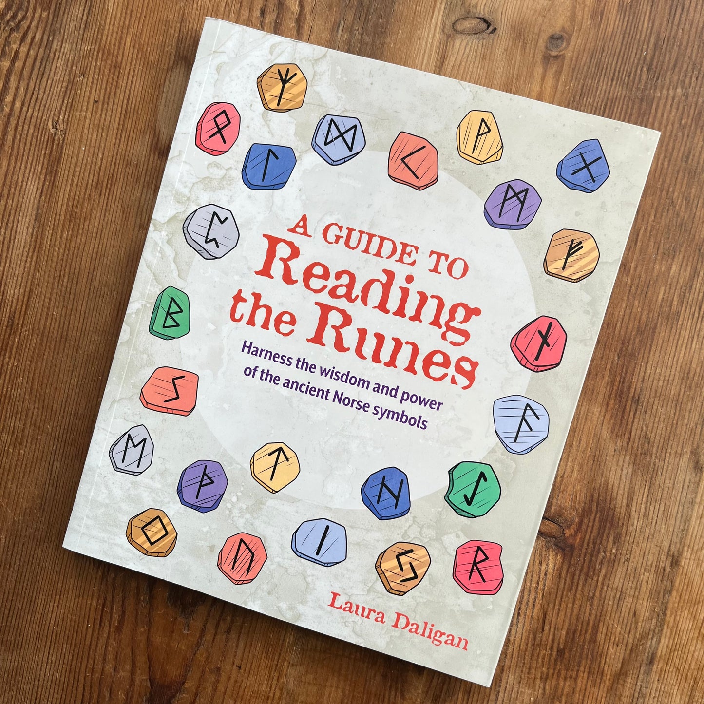 A book titled 'A Guide to Reading the Runes' by Laura Daligan, featuring a textured off-white cover decorated with colorful illustrated runic symbols, placed on a wooden surface.