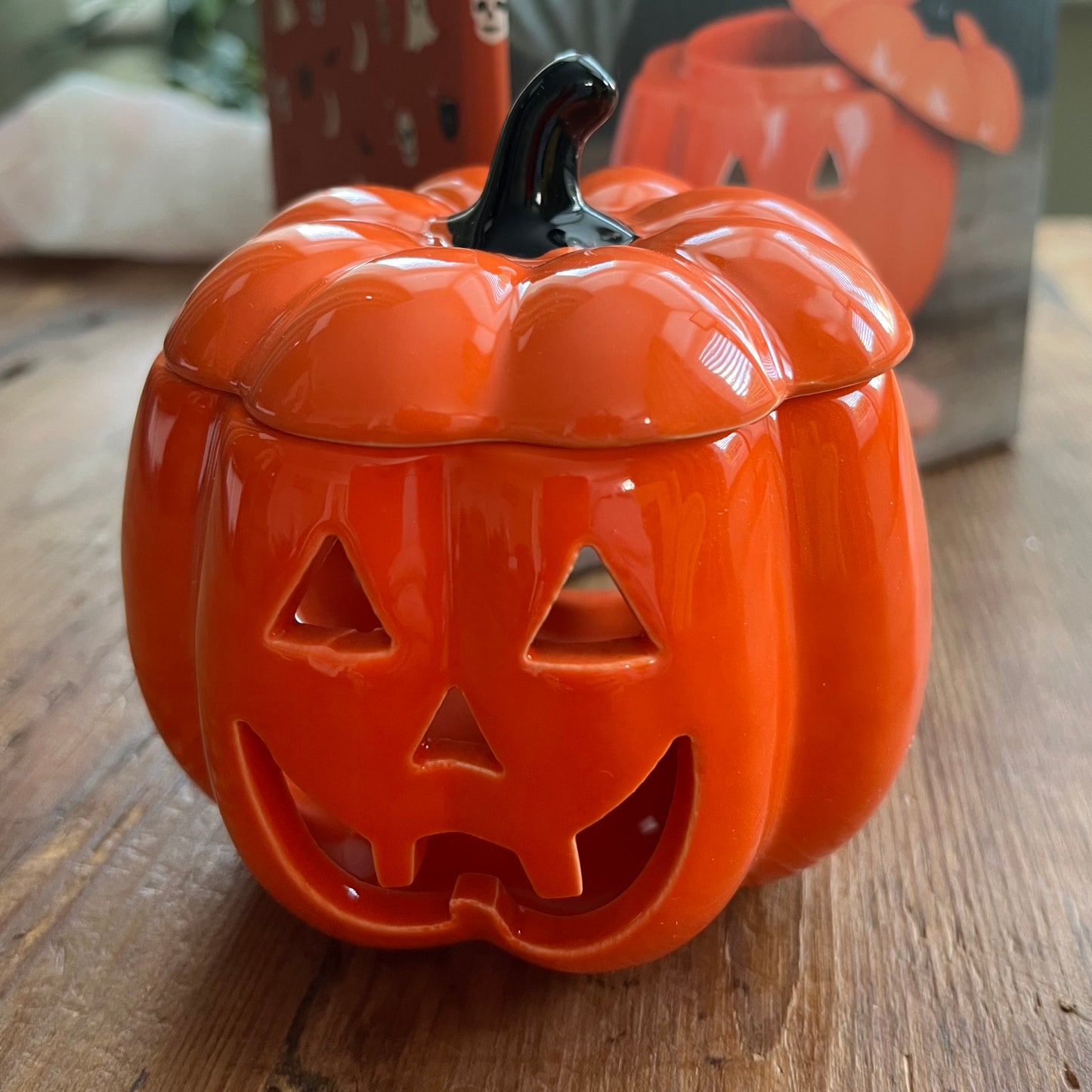 An orange pumpkin-shaped ceramic oil burner with a carved jack-o'-lantern face and a black stem, sitting on a wooden surface with a blurred background.