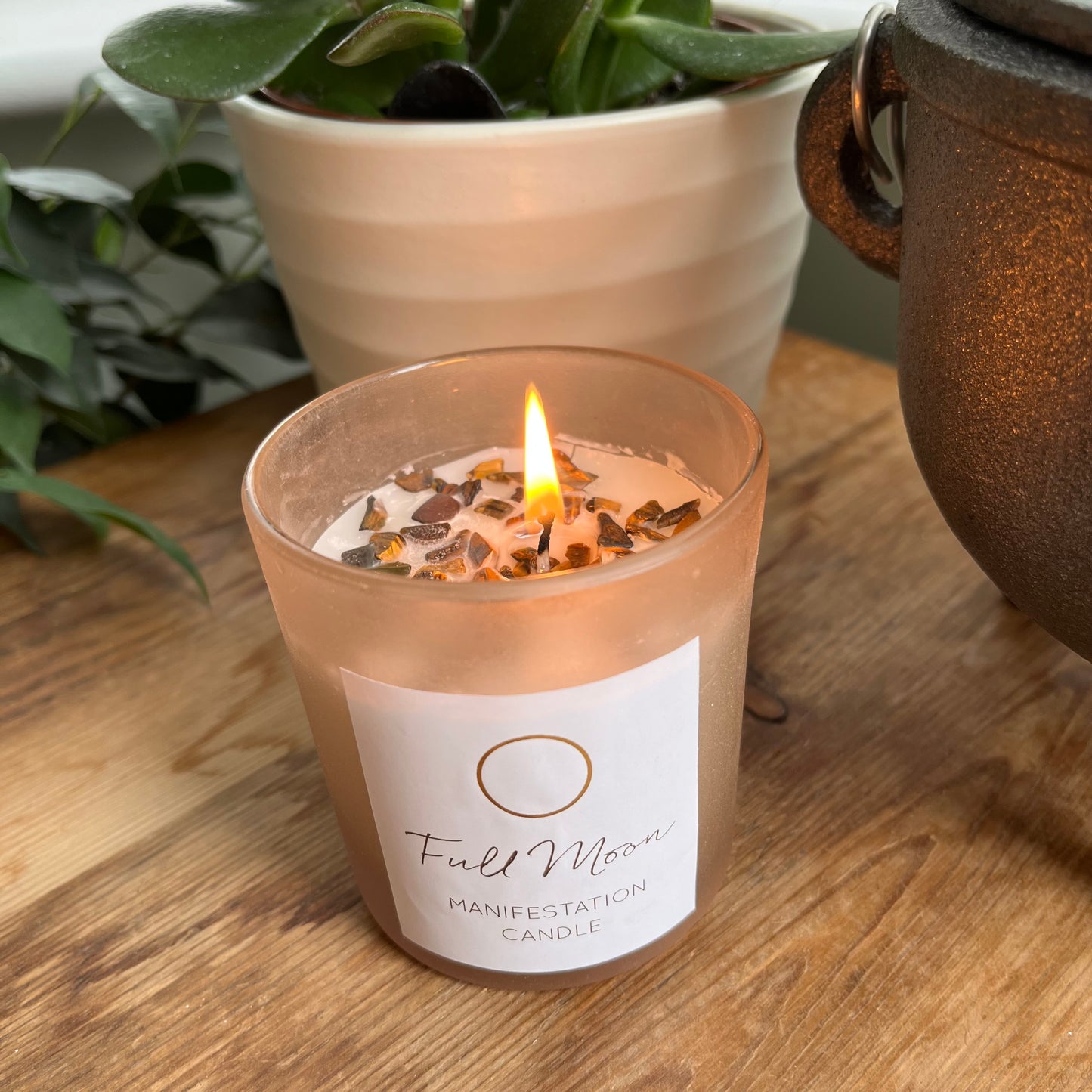 beige frosted glass candle holder containing a white candle with many Tigers eye crystals embedded in the top. it is labelled Full moon manifestation candle. The candle is lit and the candle flame creates a warm glow 