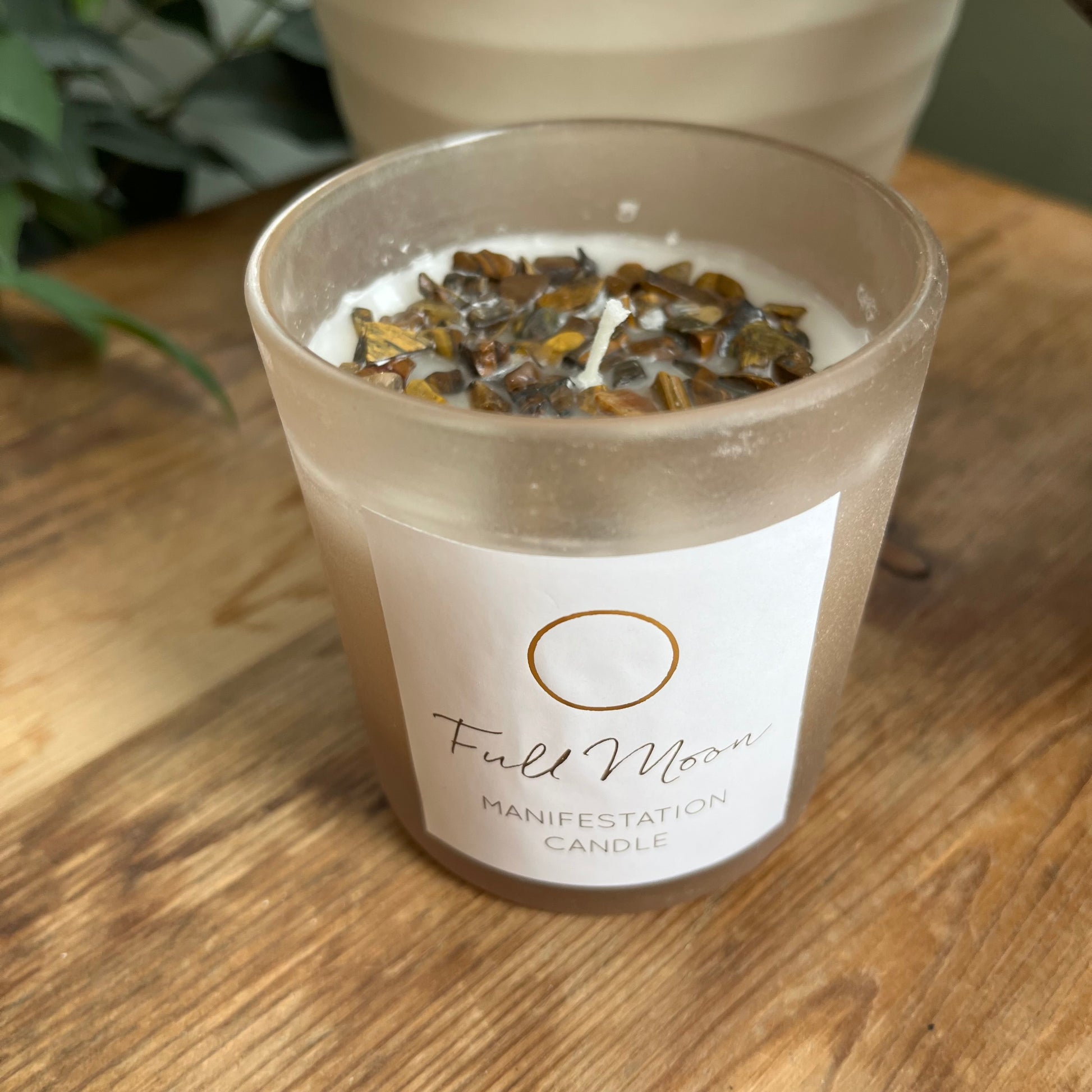 beige frosted glass candle holder containing a white candle with many Tigers eye crystals embedded in the top. it is labelled Full moon manifestation candle 