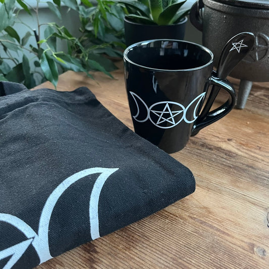 Black mug with white symbol on a wooden surface next to black fabric with similar symbol