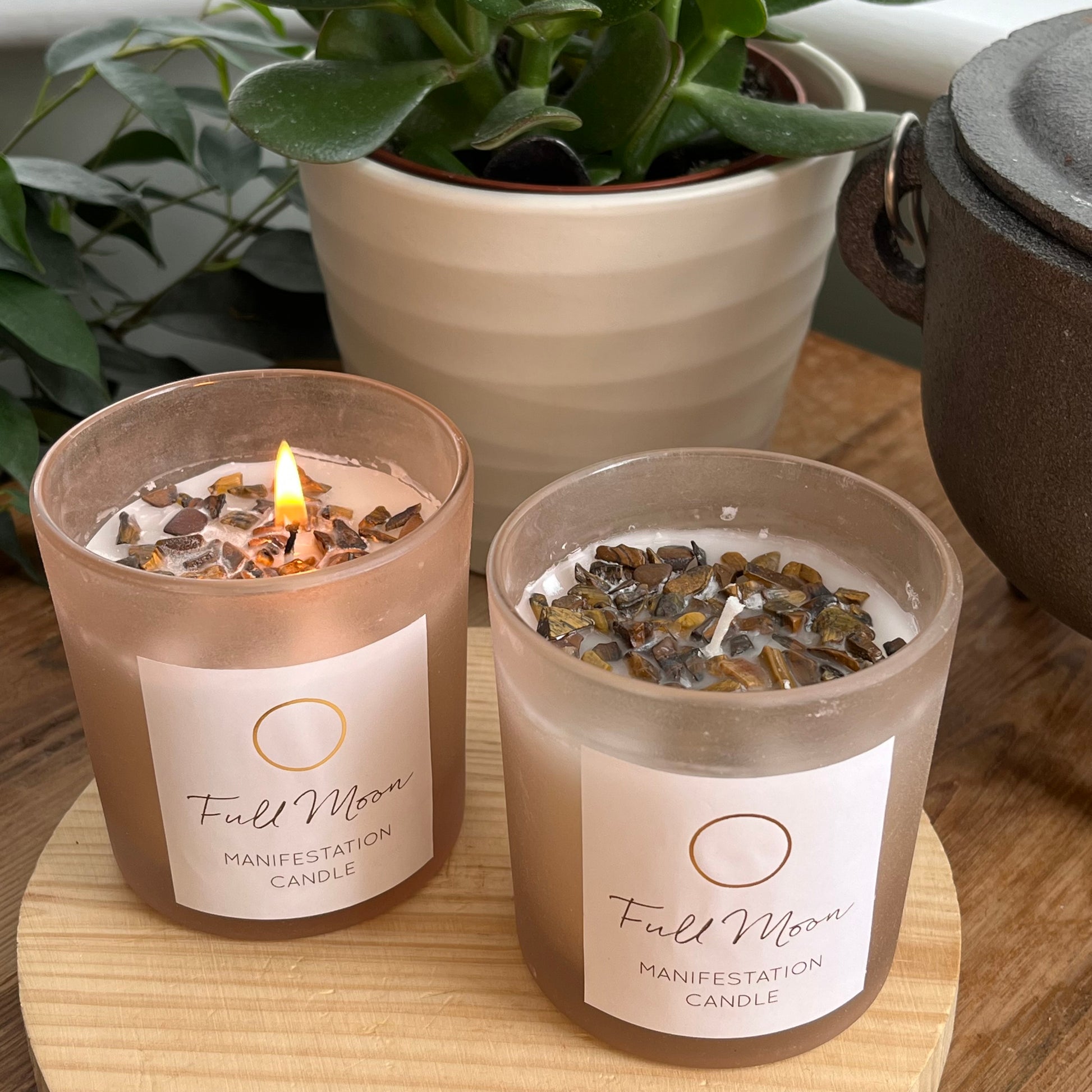 Two  Full Moon manifestation candles in beige frosted glass holder. candles are infused with Tigers eye crystals . one candle is lit and the flame creates a warm glow