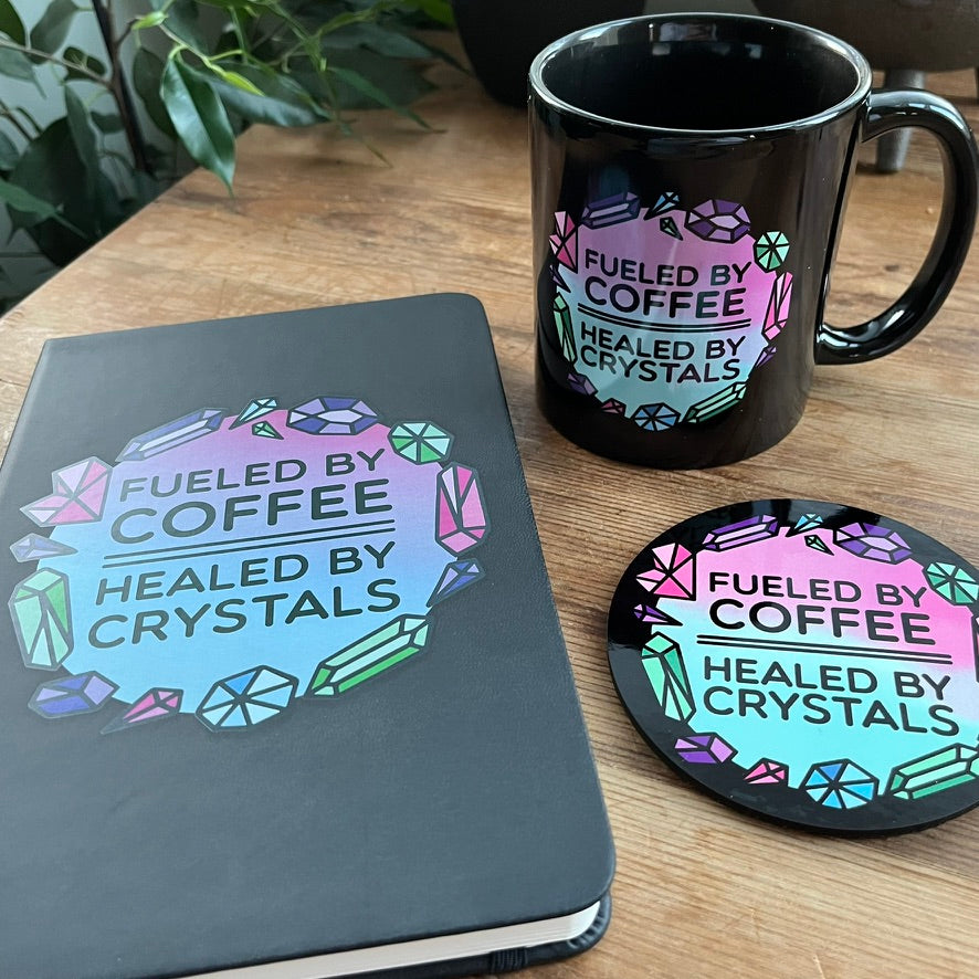 Black mug, notebook, and coaster with 'Fueled by Coffee, Healed by Crystals' design on a wooden surface.