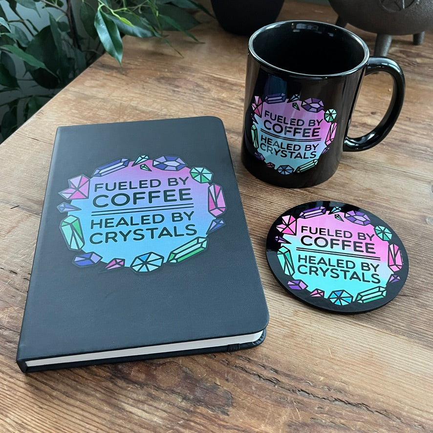 Notebook, mug, and coaster with 'Fueled by Coffee, Healed by Crystals' design on a wooden surface.