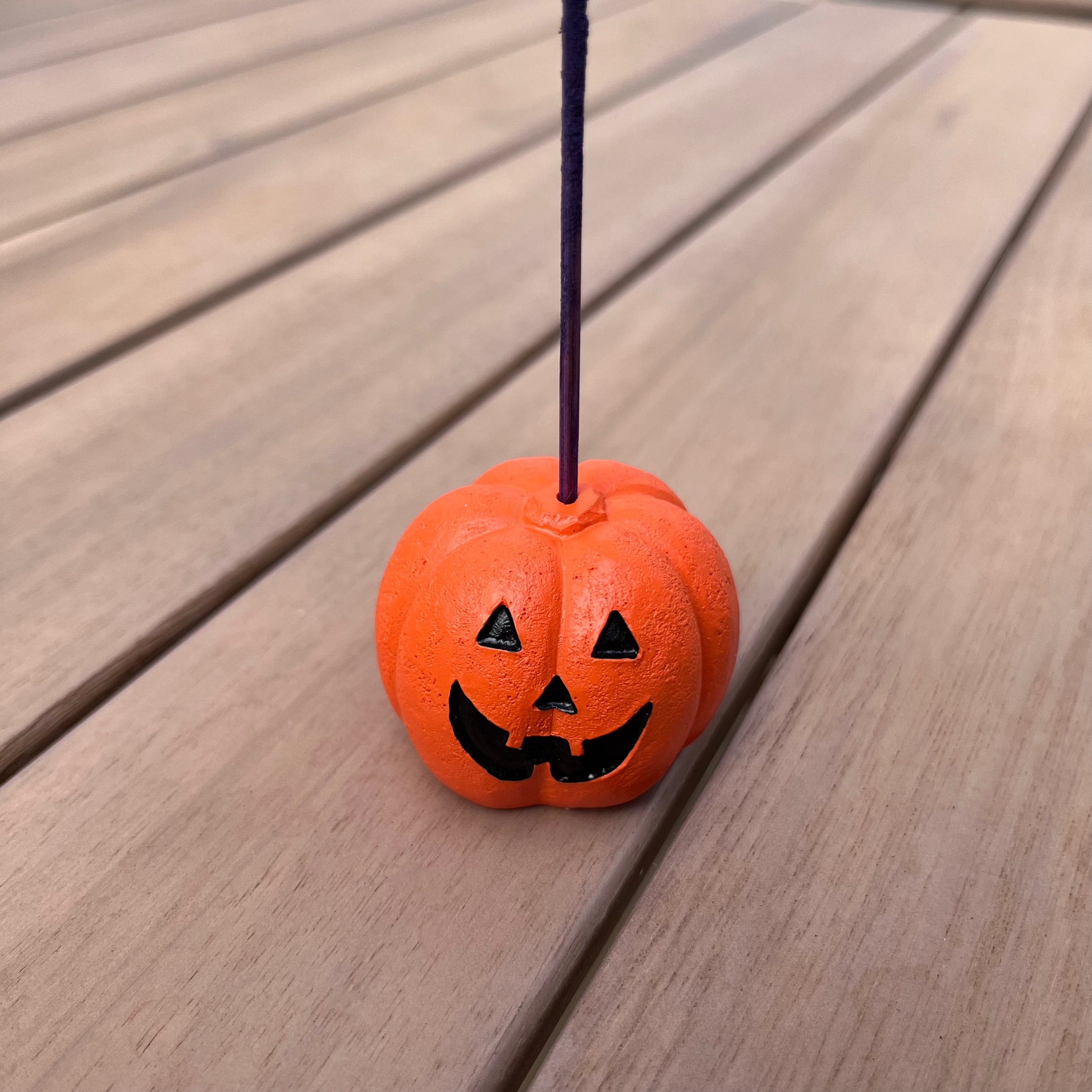 An orange pumpkin-shaped resin incense stick holder with a decorative face, placed on a wooden surface.