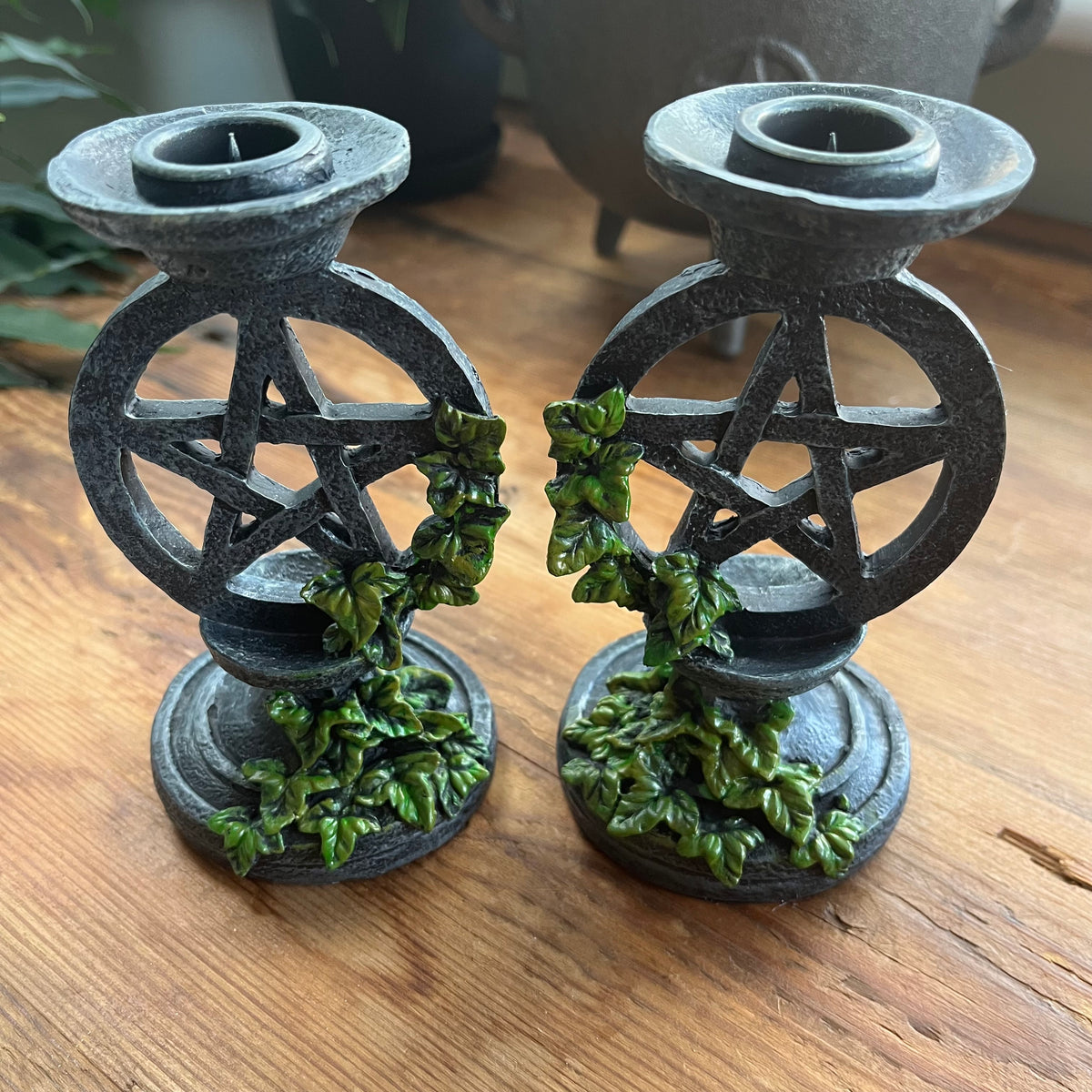 Two aged looking decorative candlestick holders featuring a pentagram design, accented with green leafy vines, shown on a wooden surface.