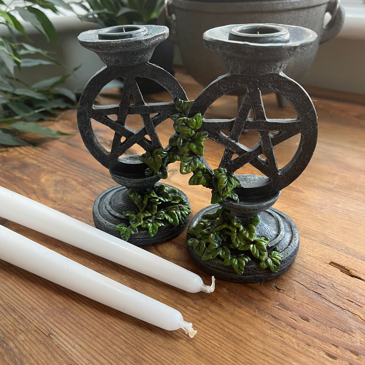 Two aged looking decorative candlestick holders featuring a pentagram design, accented with green leafy vines, shown on a wooden surface with Two white taper candles