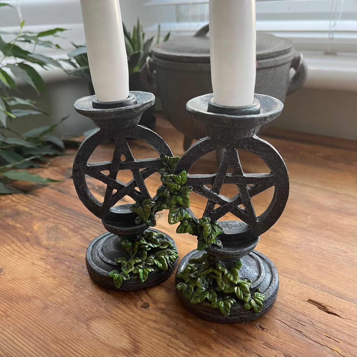 Two white taper candlesticks  held by Two aged looking decorative candlestick holders featuring a pentagram design, accented with green leafy vines, shown on a wooden surface.