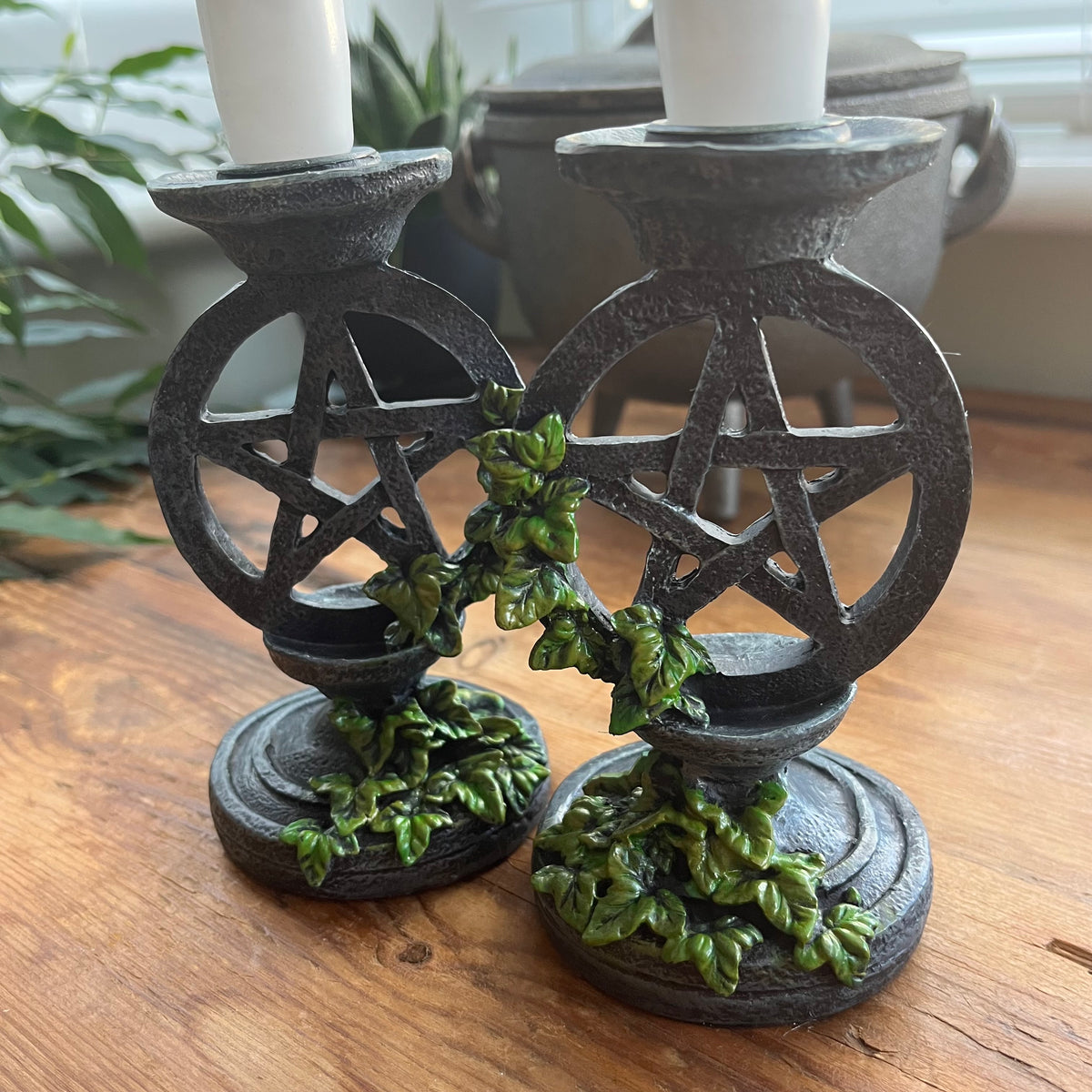 Two white taper candlesticks  held by Two aged looking decorative candlestick holders featuring a pentagram design, accented with green leafy vines, shown on a wooden surface.