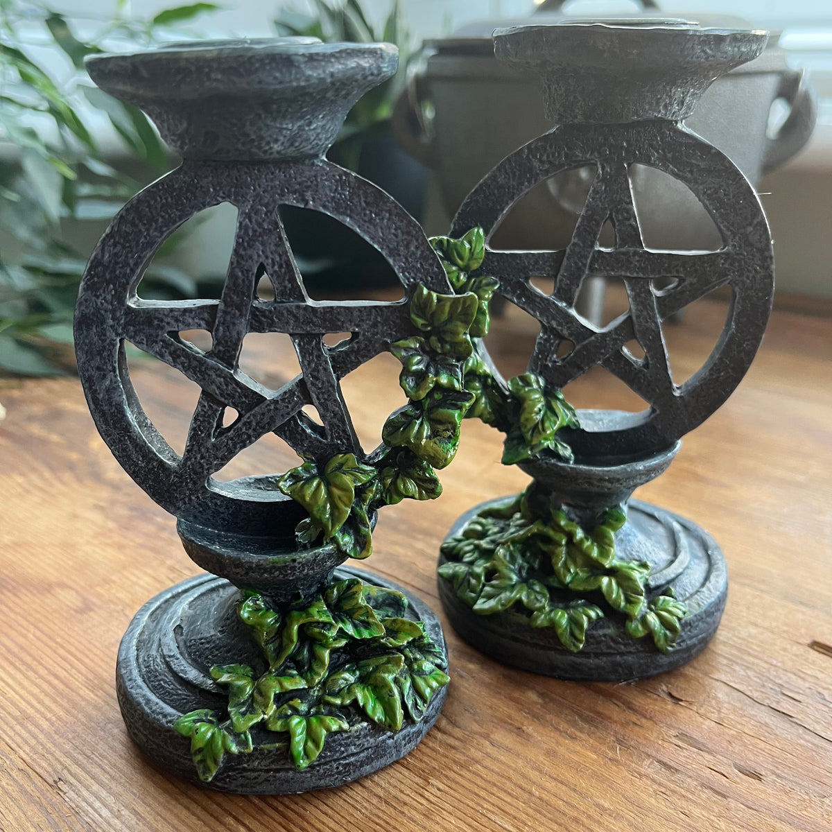  Two aged looking decorative candlestick holders featuring a pentagram design, accented with green leafy vines, shown on a wooden surface.