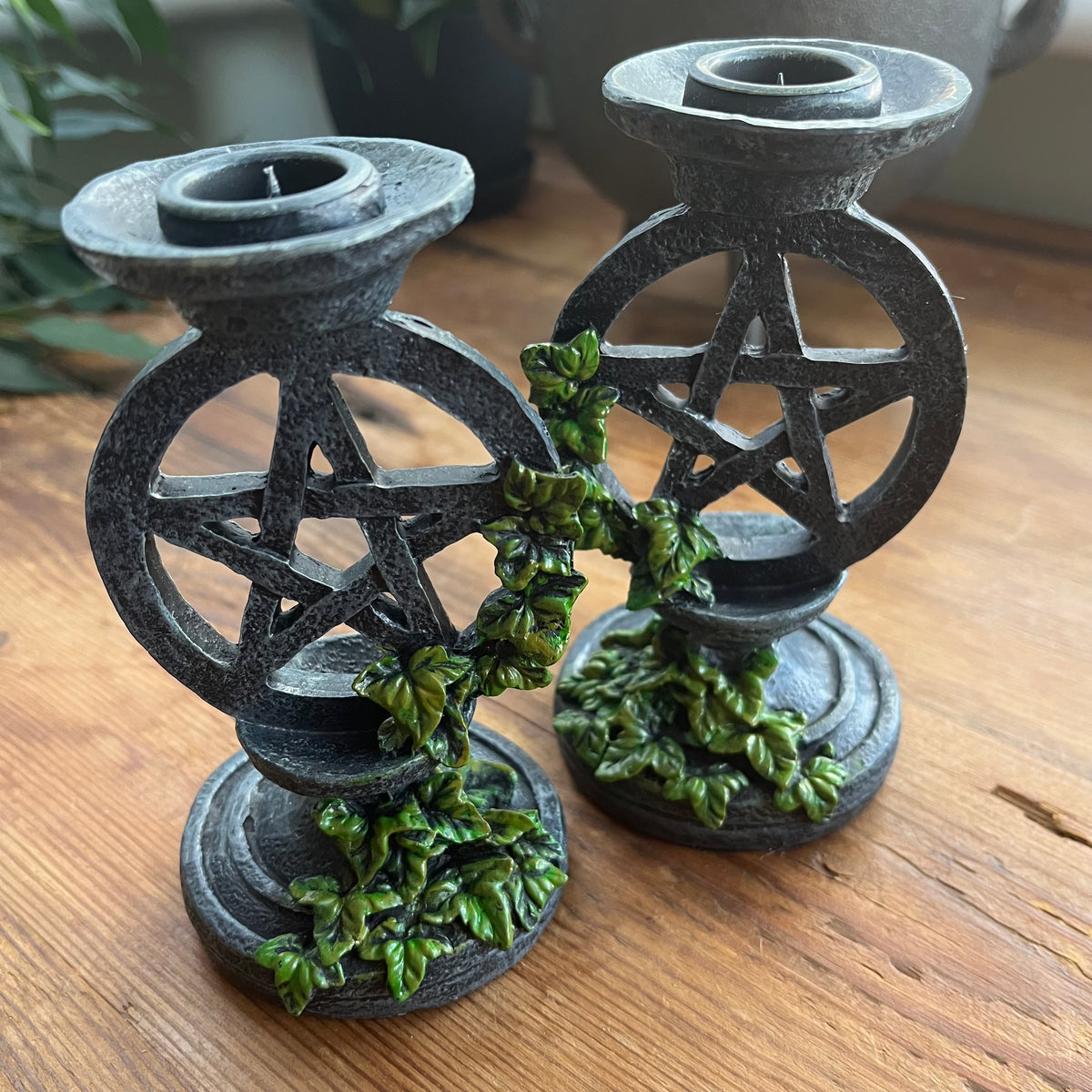  Two aged looking decorative candlestick holders featuring a pentagram design, accented with green leafy vines, shown on a wooden surface.