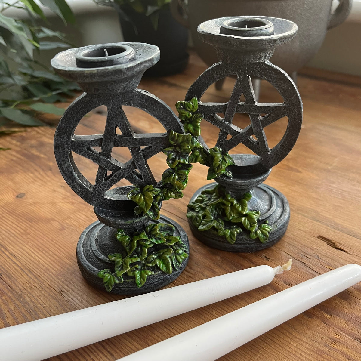  Two aged looking decorative candlestick holders featuring a pentagram design, accented with green leafy vines, shown on a wooden surface with two white taper candles