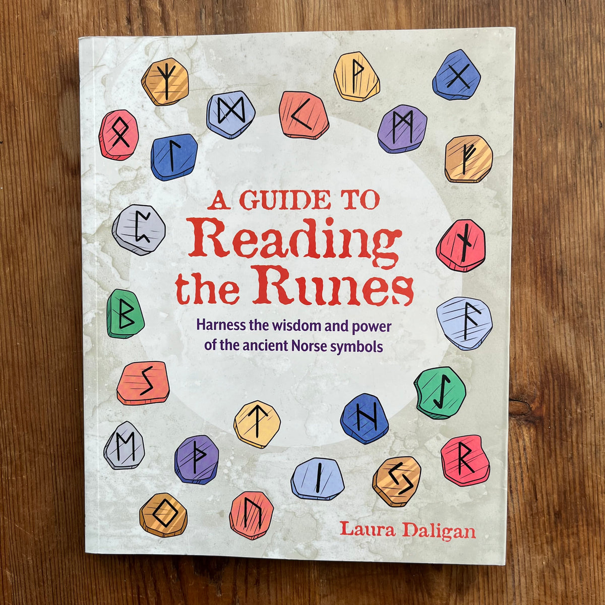 A book titled 'A Guide to Reading the Runes' by Laura Daligan, featuring a light beige cover decorated with colorful illustrated runic symbols, placed on a wooden surface.