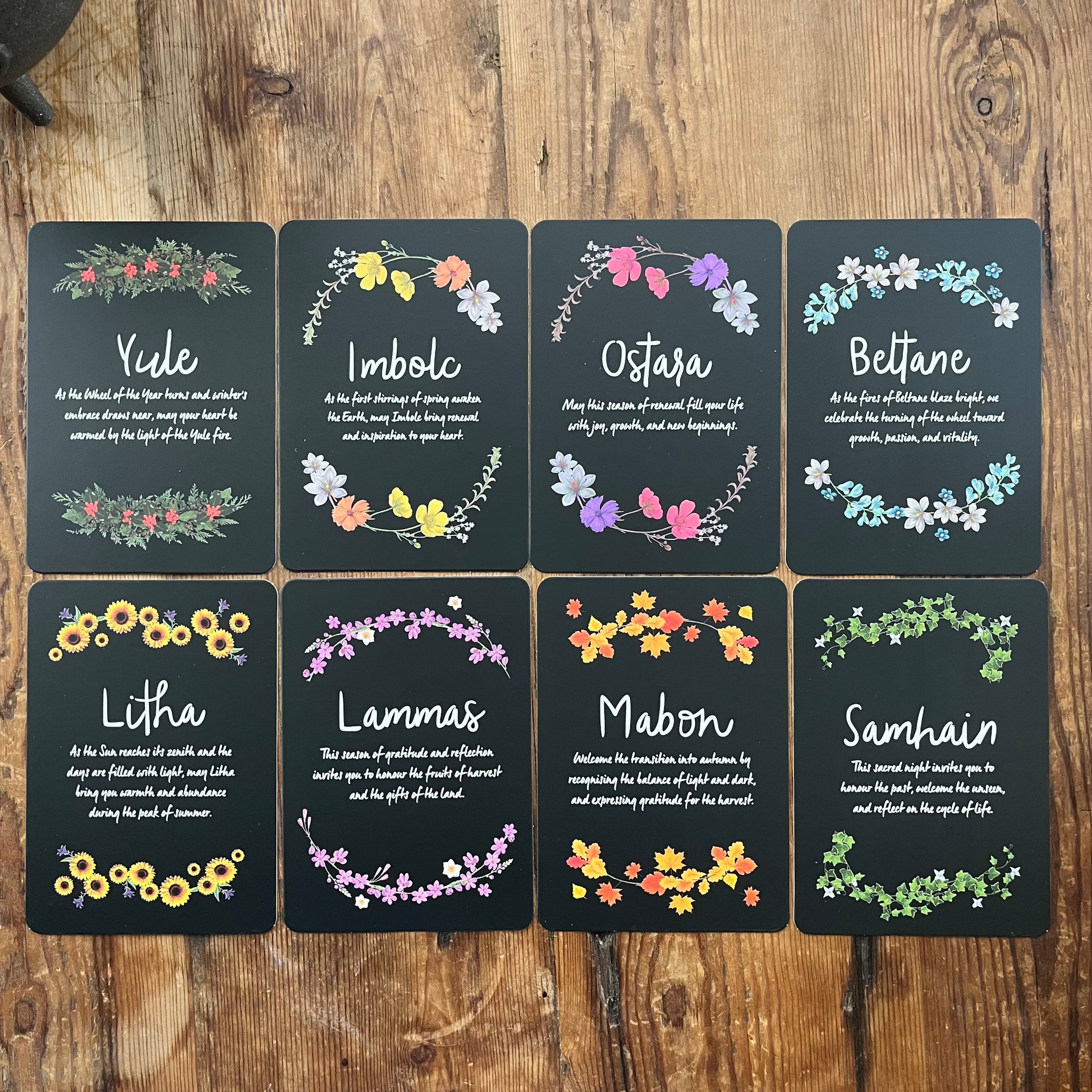 Set of eight cards with floral designs and Sabbat names on a wooden surface