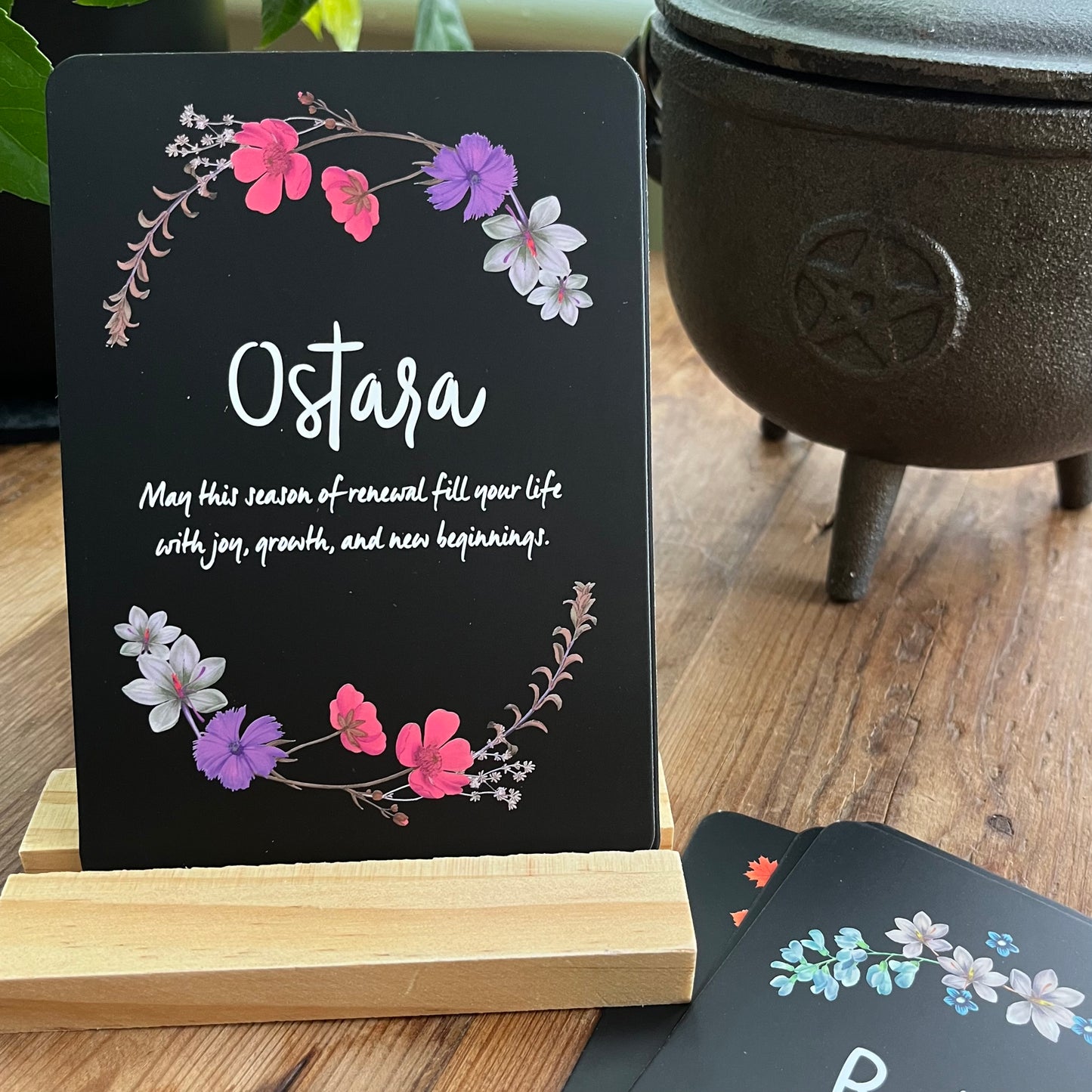 Decorative card with floral design and 'Ostara' text on a stand, with a black pot in the background.