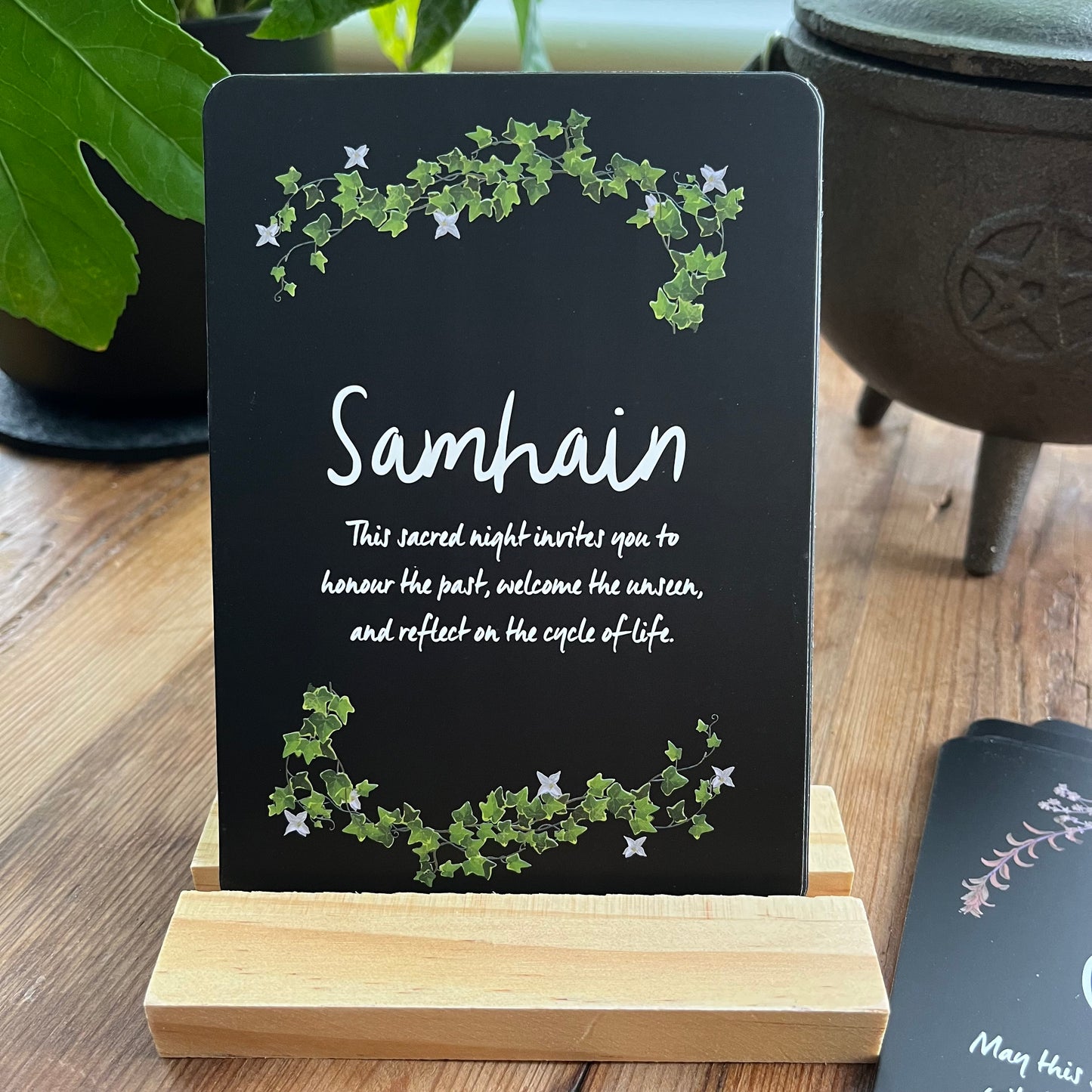 Black sign with 'Samhain' text and decorative elements on a wooden stand, with a pot and plant in the background.