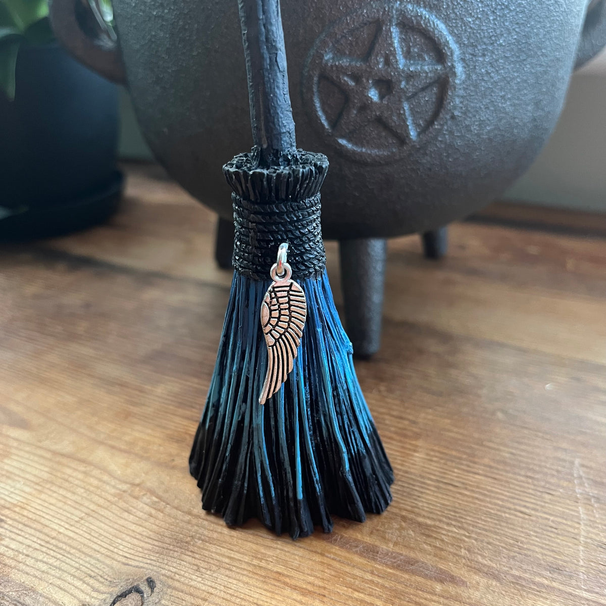 A close-up of a black and blue tassel broom with a Angel wing charm, standing on a wooden surface in front of a dark cauldron.