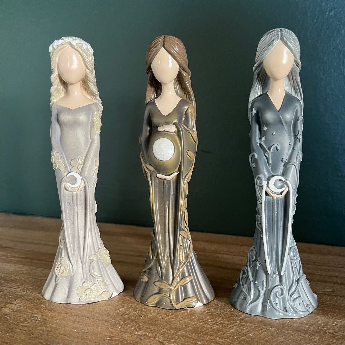 Three decorative figurines of pregnant goddesses in long flowing robes, standing on a wooden surface against a dark teal background.