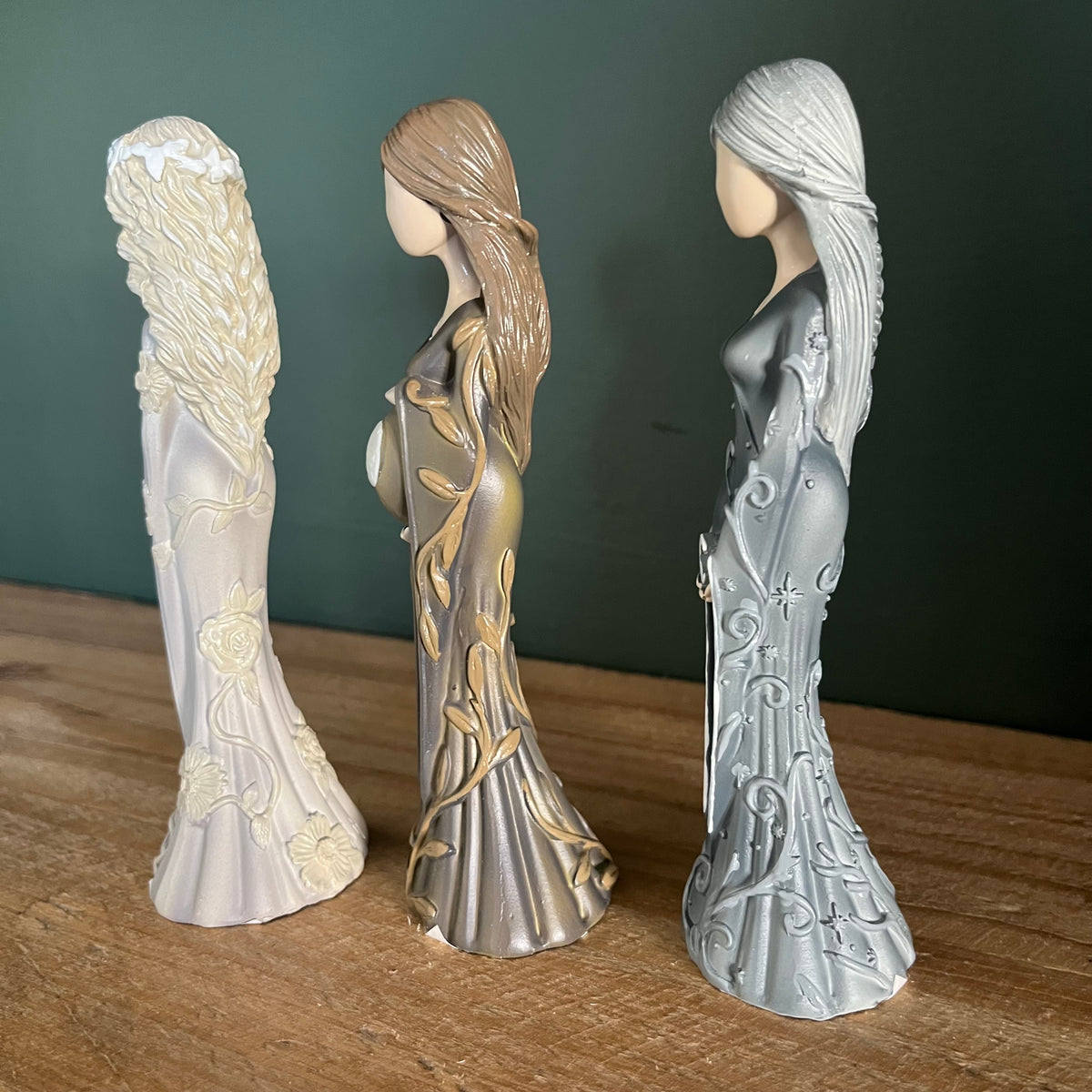 Three decorative female figurines in profile standing on a wooden surface against a dark green background. 
