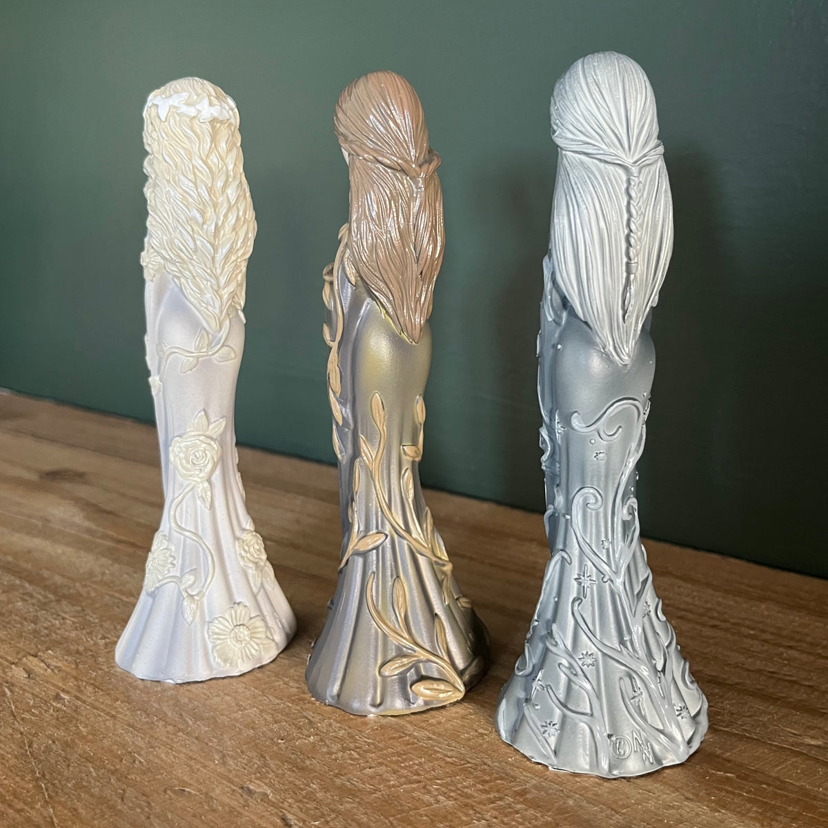 Three decorative figurines with long flowing hair and floral vine details, shown from the back on a wooden surface against a dark green wall.
