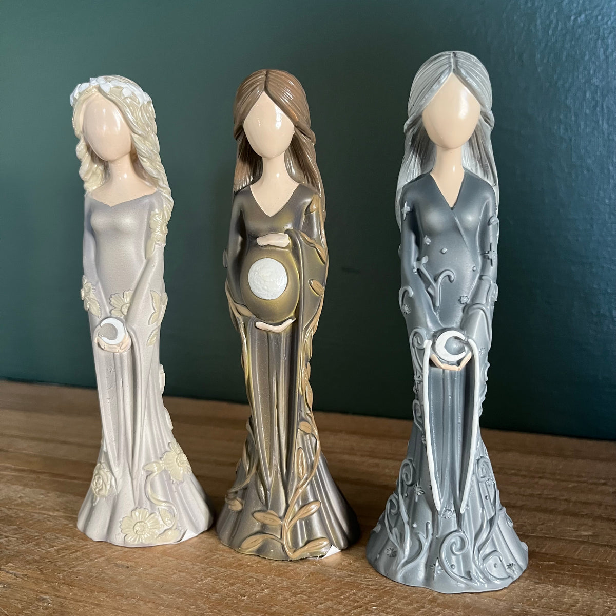 Three decorative figurines of pregnant goddesses standing side by side on a wooden surface against a dark teal background. The left figurine is light gray with cream accents, the middle is gray and gold with a white belly emblem, and the right is metallic silver with a white belly emblem.