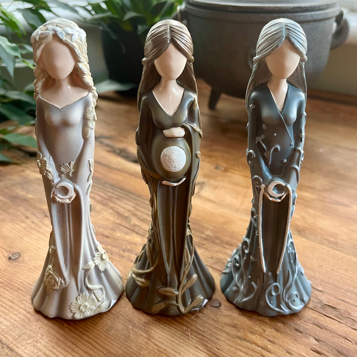 Three decorative figurines of a pregnant women in long flowing robes, displayed on a wooden surface with a potted plant and pot in the background.