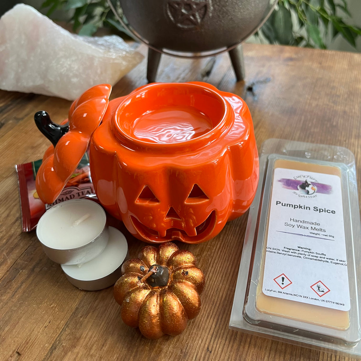 A Halloween-themed product arrangement on a wooden surface featuring a bright orange pumpkin-shaped wax melt burner, a package of Pumpkin Spice handcrafted soy wax melts, white tea lights, a small metallic gold pumpkin ornament