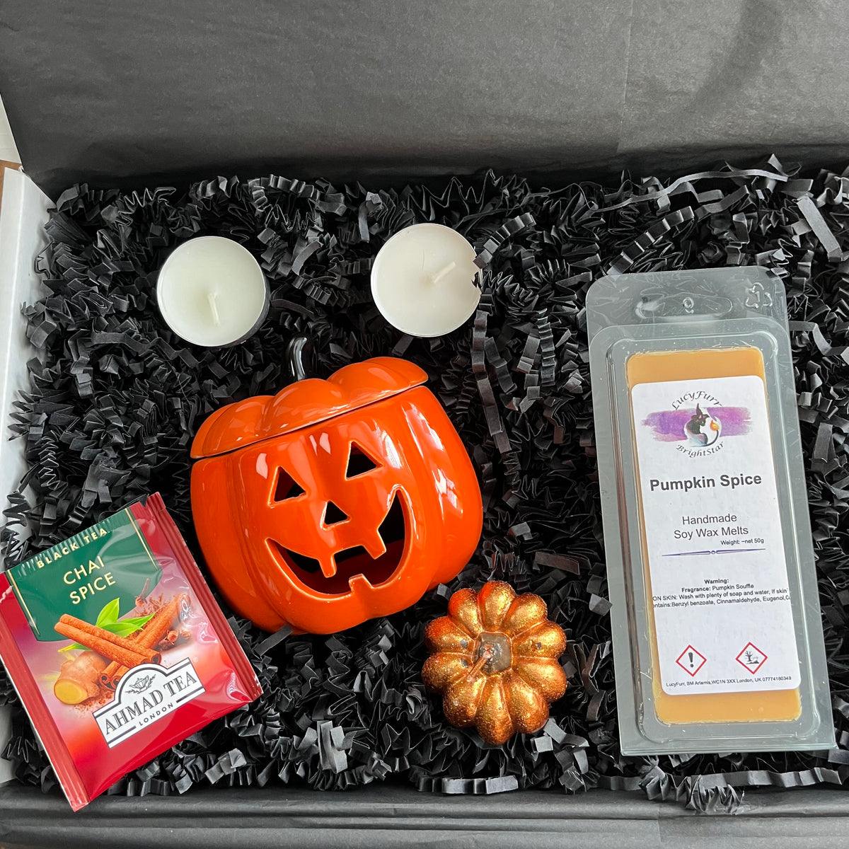 A curated gift box set featuring a bright orange jack-o'-lantern ceramic oil burner a package of Almad Tea Chai Spice, and pumpkin spice soy wax melts, arranged in black shredded paper inside a  box with two white tea lights.