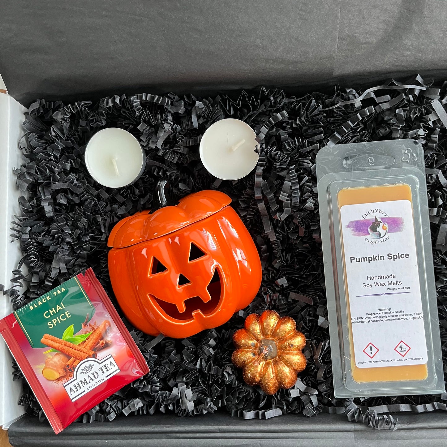 A curated gift box set in black shredded paper featuring a bright orange pumpkin-shaped ceramic oil burner a package of 'Pumpkin Spice' soy wax melts a packet of Ahmed Tea 'Cinnamon Spice' tea, and two white tea light candles and a orange pumpkin shaped candle