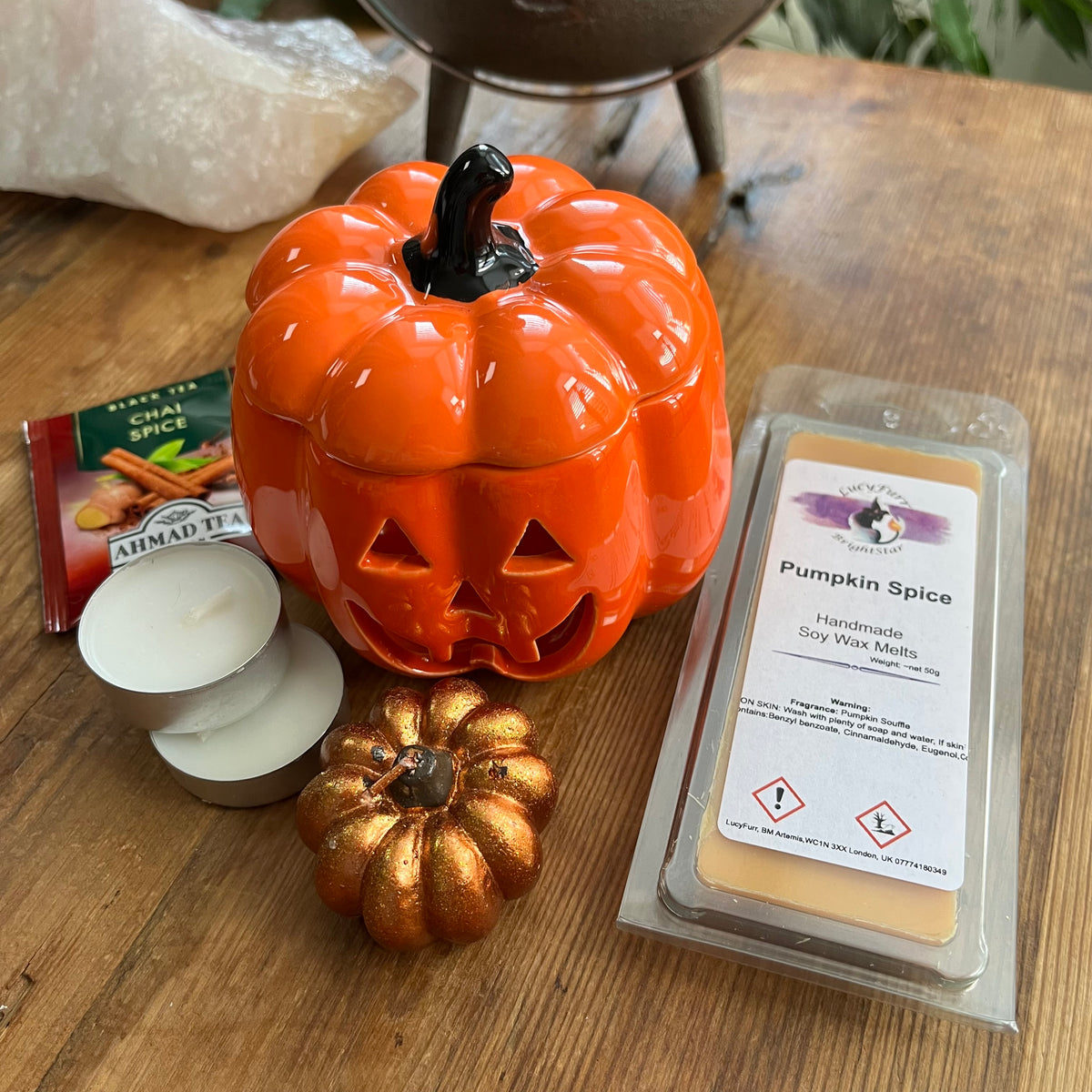 A styled tabletop arrangement featuring a glossy orange jack-o'-lantern ceramic oil burner, a small pumpkin shaped candle, a package of Ahmad Tea spice tea packet, two white tea light candles, and a packaged wax melt labeled 'Pumpkin Spice' on a wooden surface.