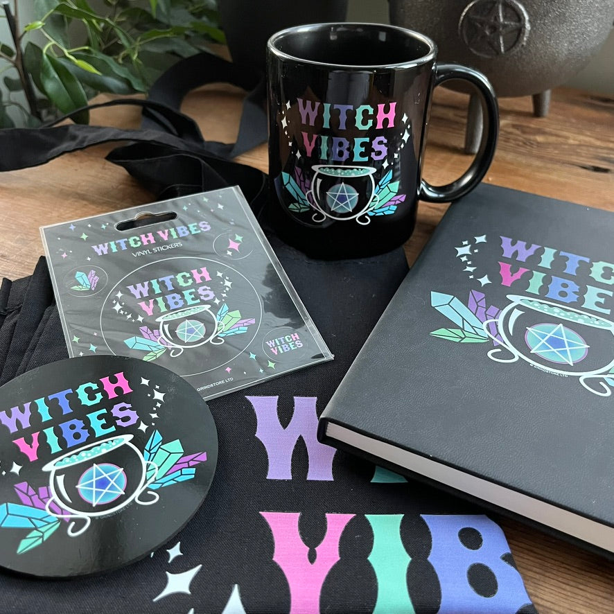 Black mug, notebook, coaster, and stickers with 'Witch Vibes' design on a wooden surface.
