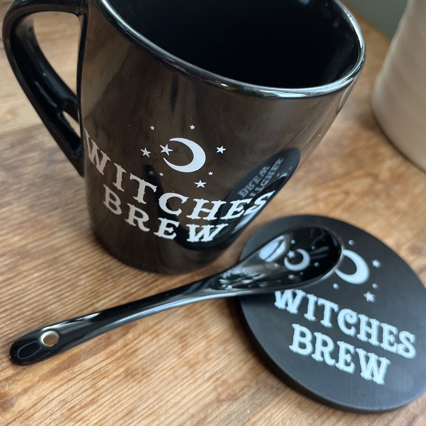 Black mug with the words Witches brew and a moon and star design in white. A matching spoon sits on a coaster that also features the words witches brew in the same design 