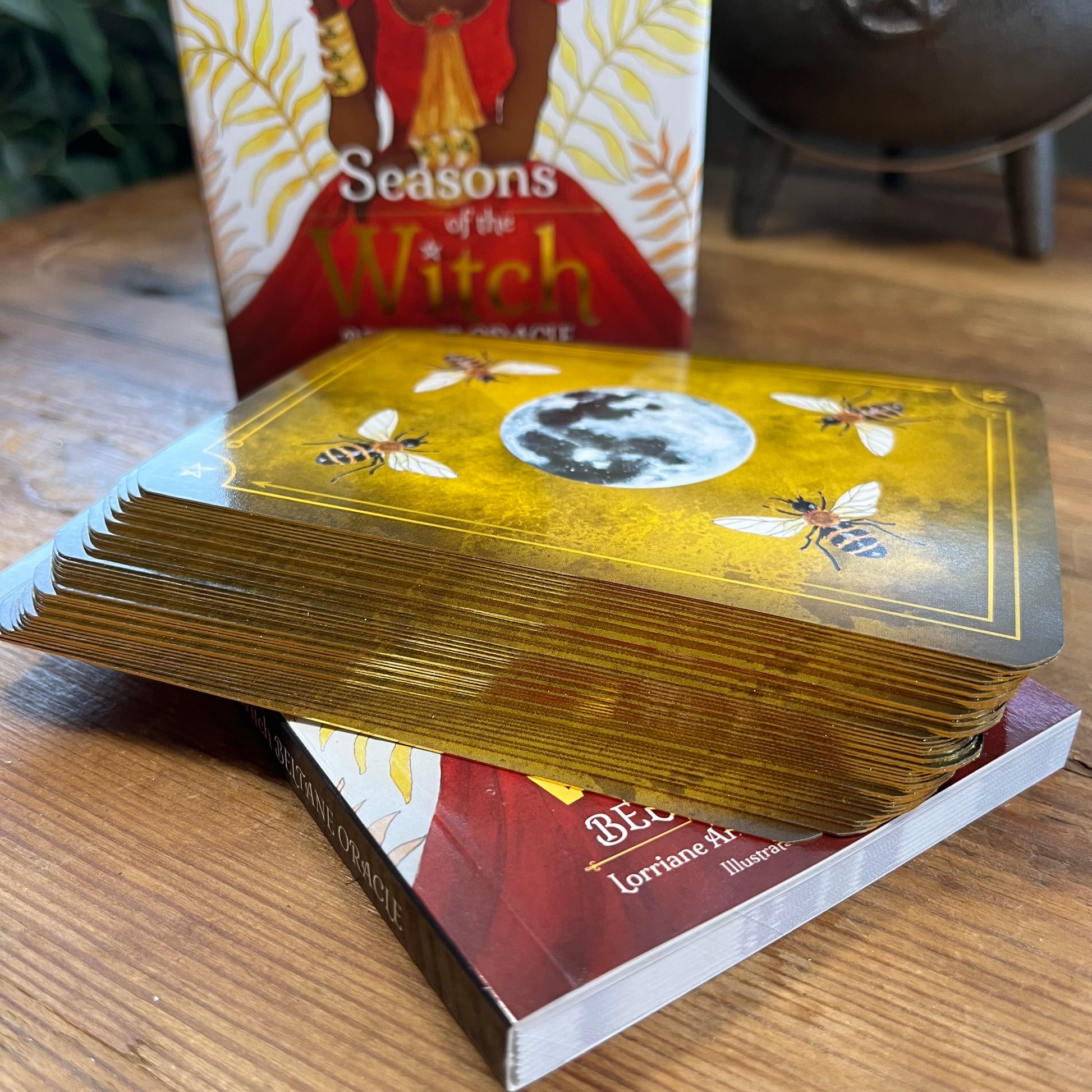 Stack of Oracle cards with bee and moon design on a wooden surface, with 'Seasons of the Witch' book in the background.