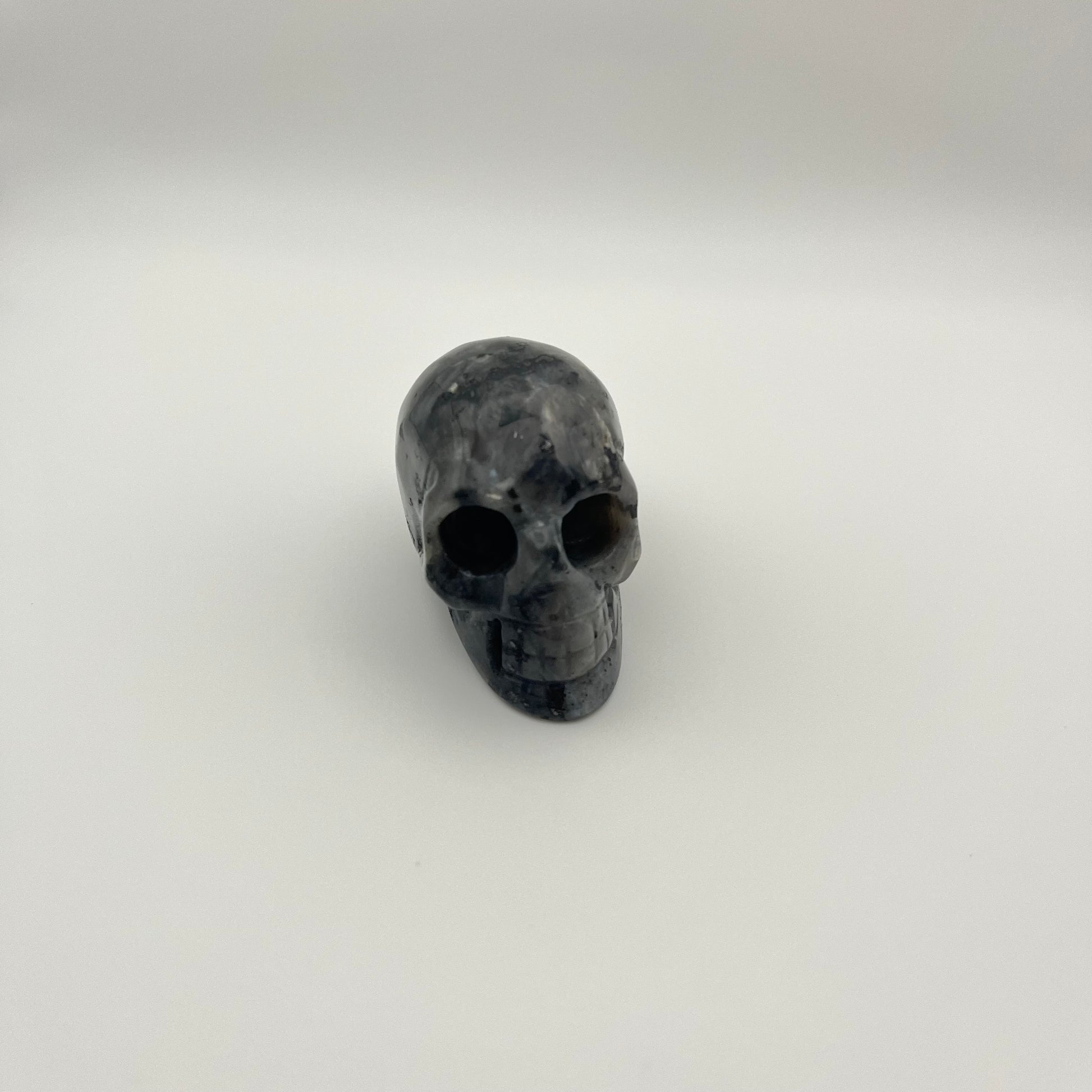 A small carved skull figurine made of dark polished stone, likely obsidian, with natural marbled gray and black patterns, set against a plain light gray background.