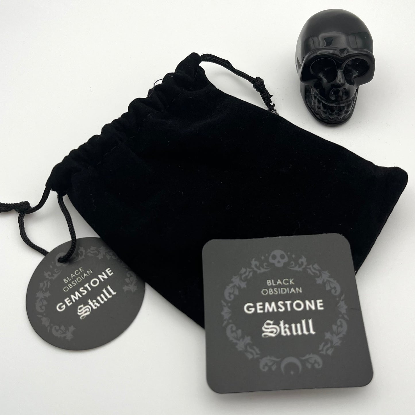 Black obsidian gemstone skull displayed with a black velvet drawstring pouch, a round black tag, and a square product card on a white background.