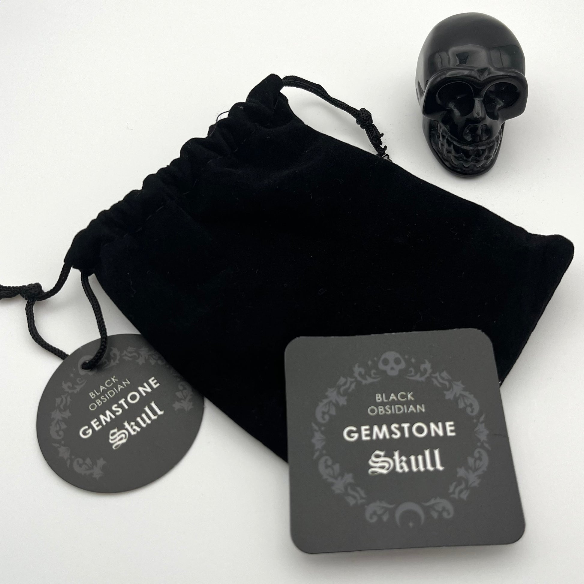 Black obsidian gemstone skull displayed with a black velvet drawstring pouch, a round black tag, and a square product card on a white background.