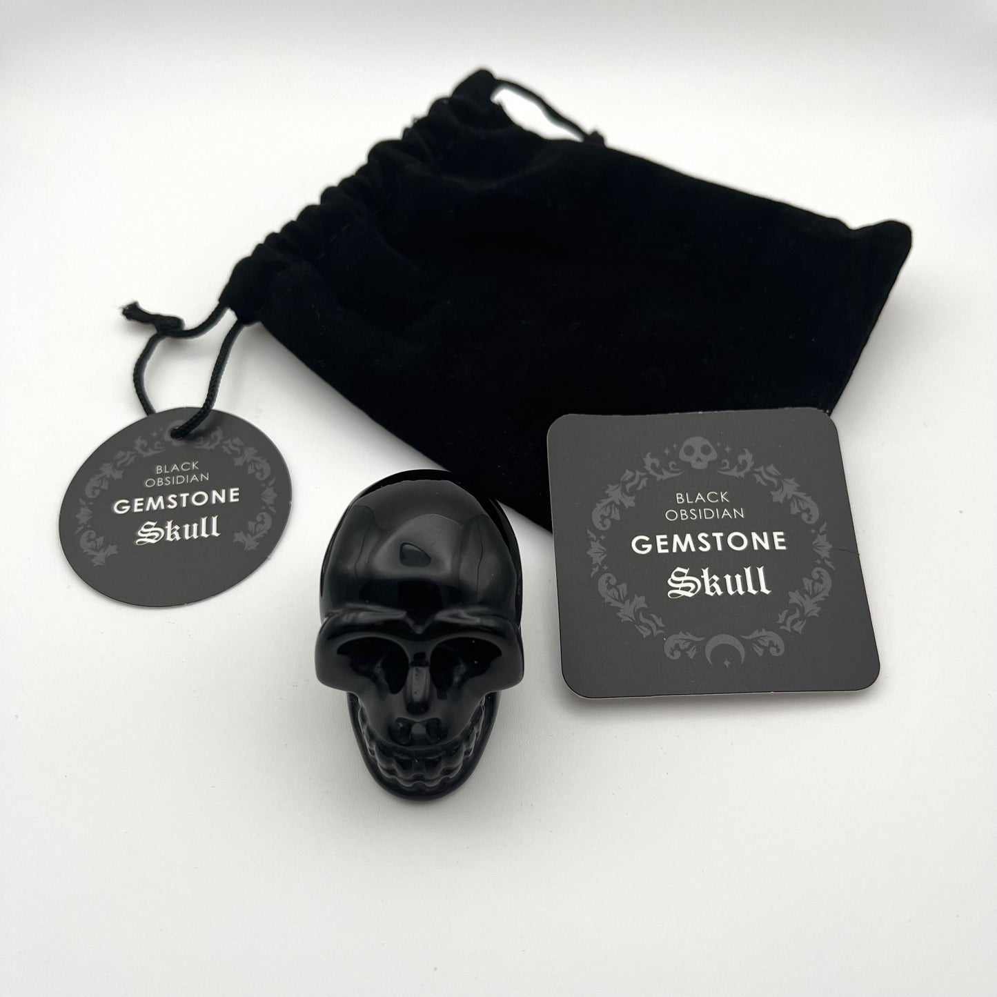A black gemstone skull figurine displayed with its black velvet drawstring pouch, a round hanging tag, and a square card on a white background. The tags and card are labeled 'BLACK OSSIDIAN GEMSTONE Skull'.