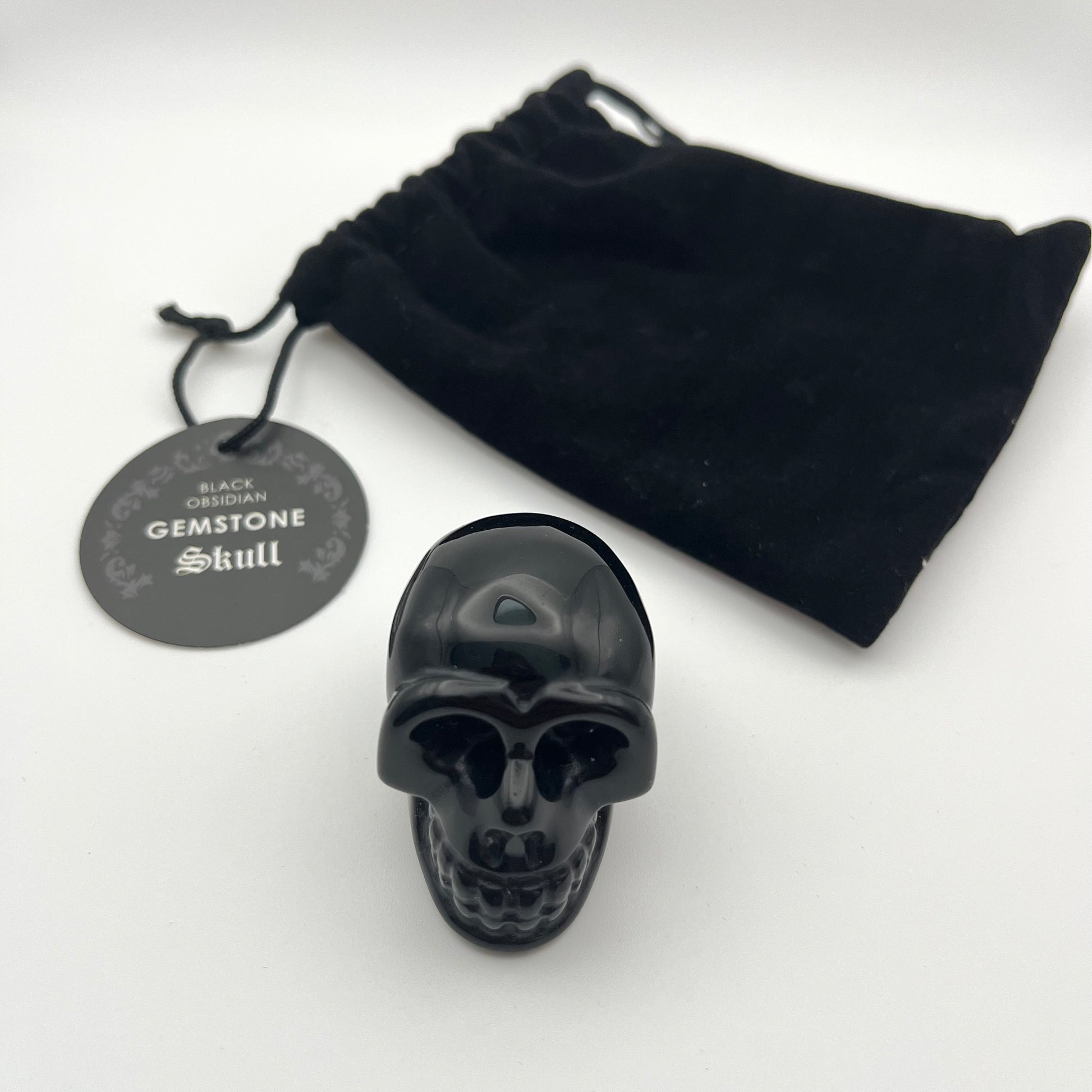 A glossy black skull-shaped gemstone with a black velvet drawstring pouch and a round black hangtag reading 'Black Obsidian Gemstone Skull' on a white background.