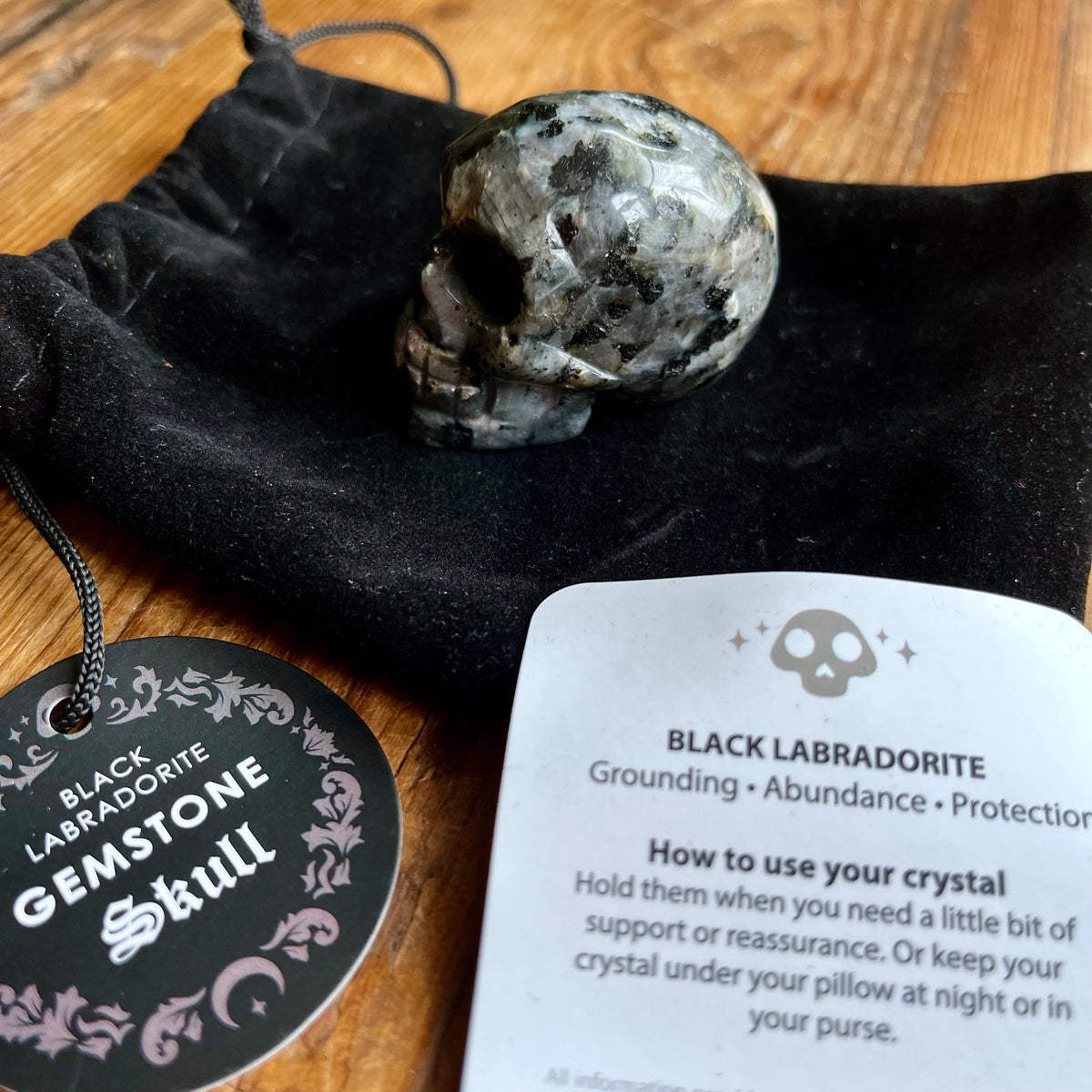 A black labradorite gemstone skull crystal resting on a black velvet drawstring pouch, with a round black tag reading 'Black Labradorite Gemstone Skull' and an informational card showing crystal usage, placed on a wooden surface.