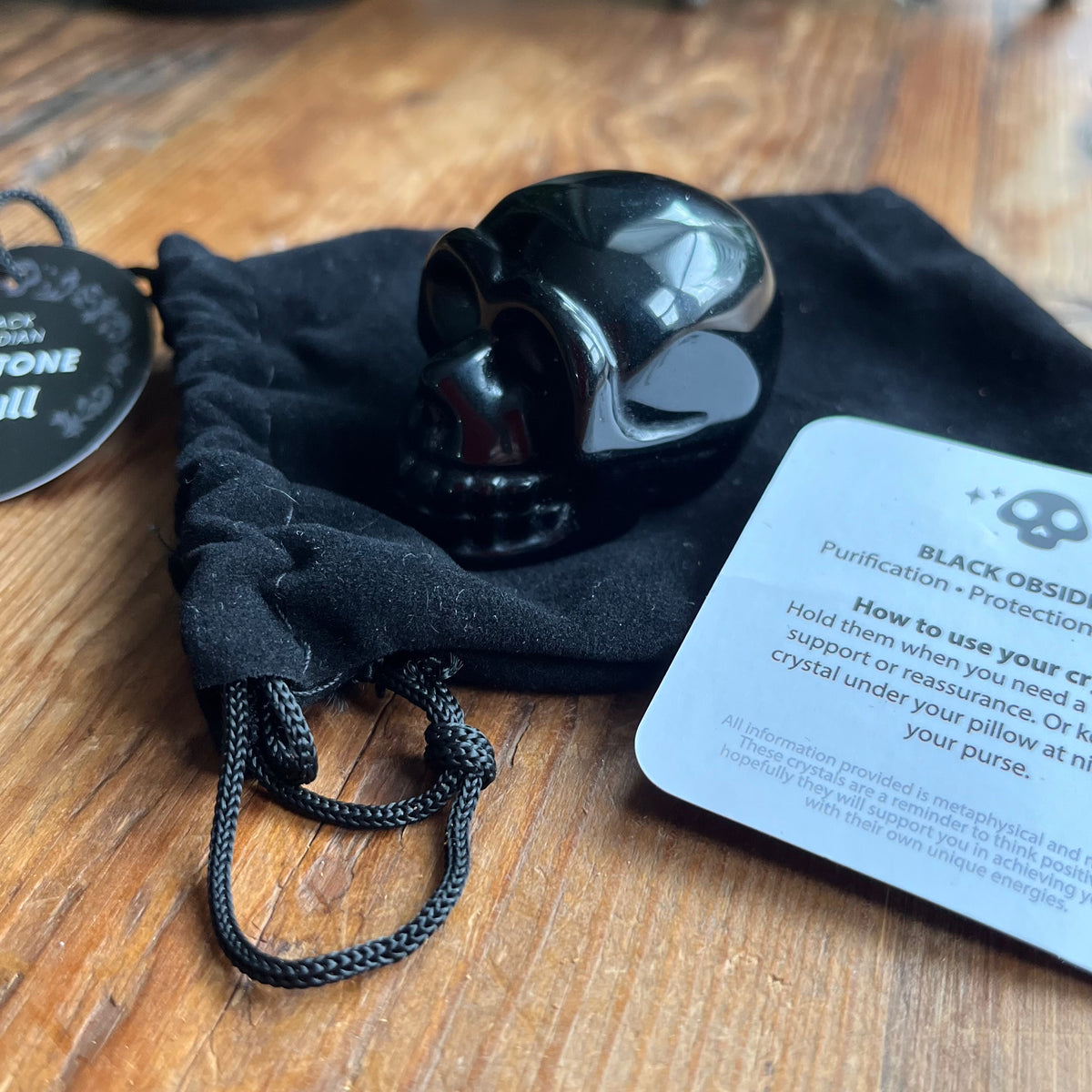 A glossy black obsidian skull resting on a black velvet drawstring pouch, with a product tag and an instruction card on a wooden tabletop.