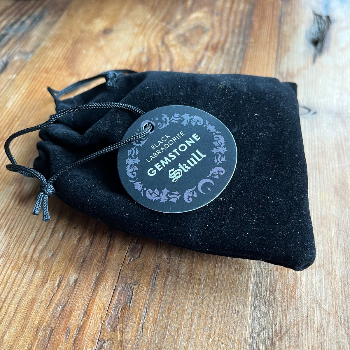 A black velvet drawstring pouch with a round black hangtag reading 'Black Lab Co. Gemstone Still' in white and lavender, placed on a rustic wooden surface.
