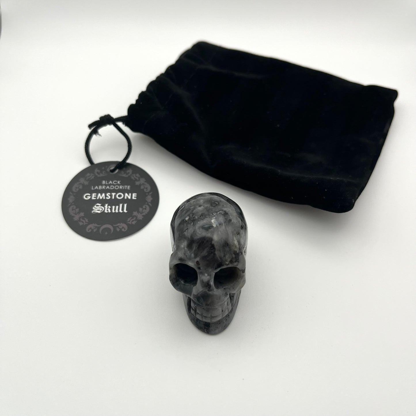 A small black gemstone skull figurine displayed on a white surface next to its black velvet drawstring pouch and a round black tag that reads 'Black Labracadore Gemstone Skull'.